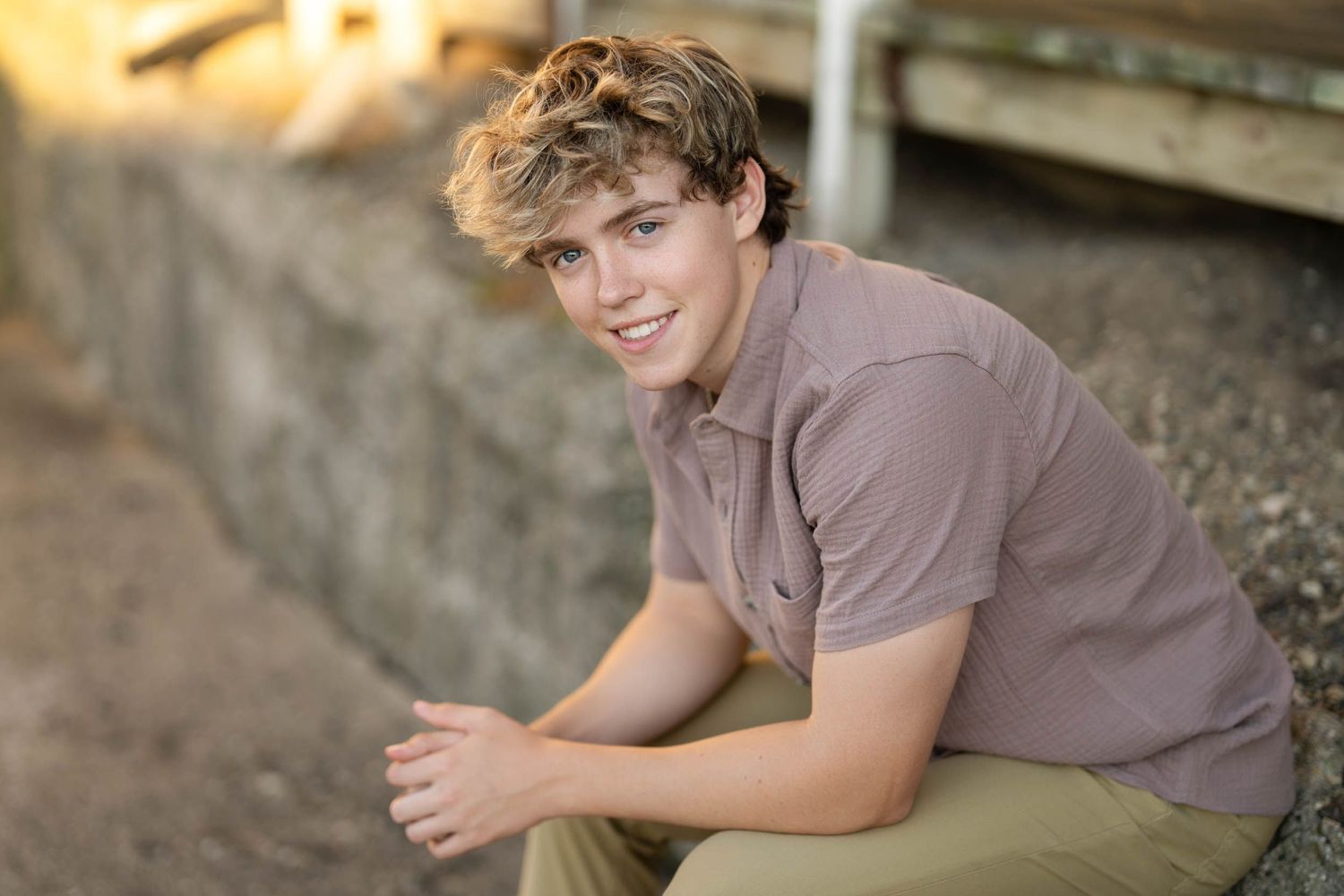 A person in a gray shirt smiles while sitting outdoors near a stone wall in natural lighting.