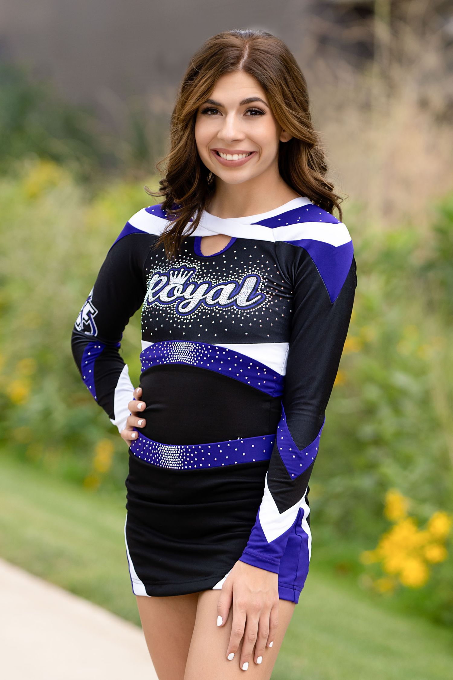 Cheerleader poses in black and blue uniform against natural outdoor backdrop.