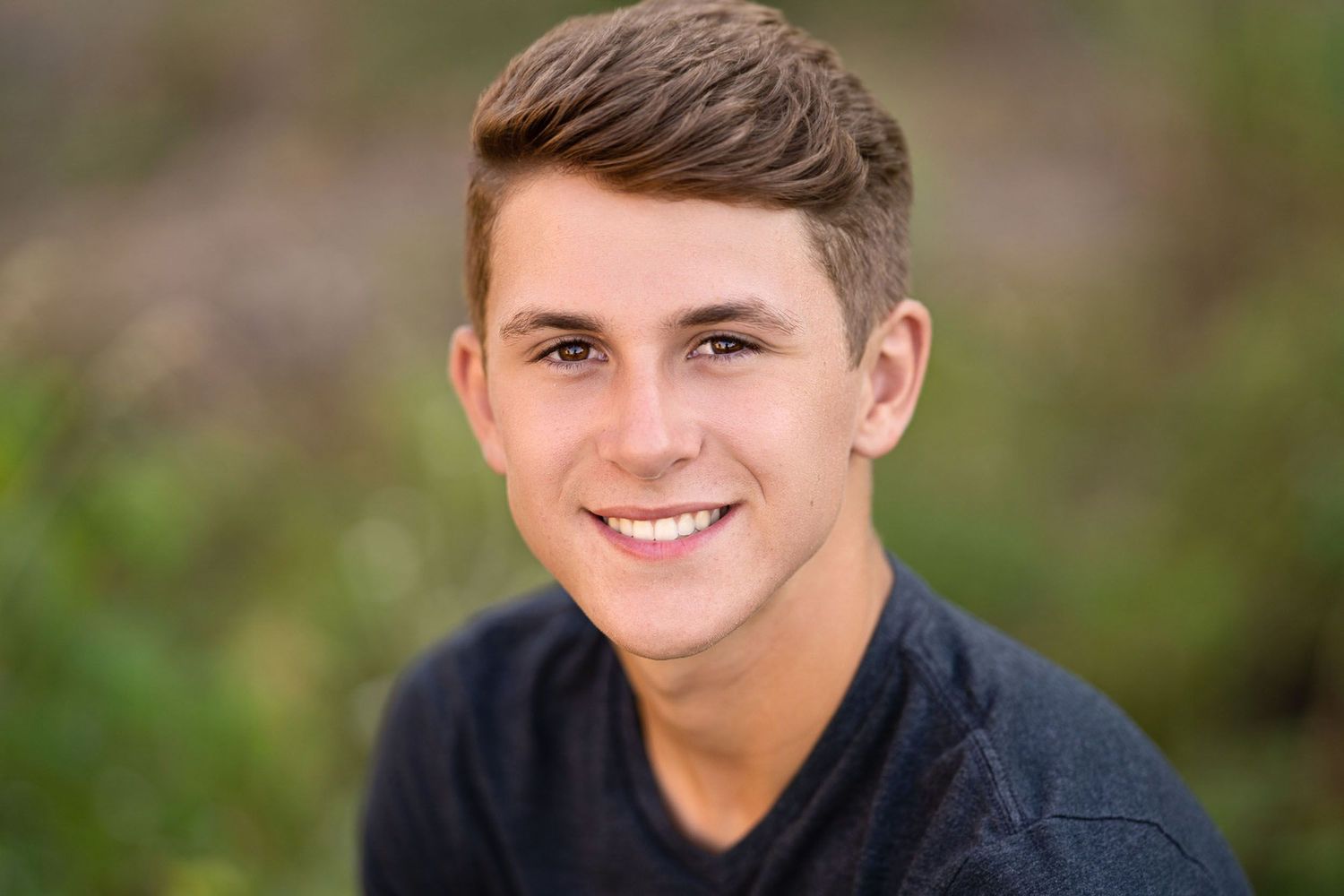A person wearing a gray sweater smiles warmly while posing outdoors against a blurred natural background.