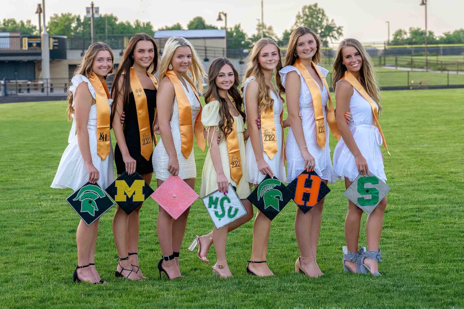 A group in white dresses and yellow sashes hold decorated graduation caps while standing on a grassy field.