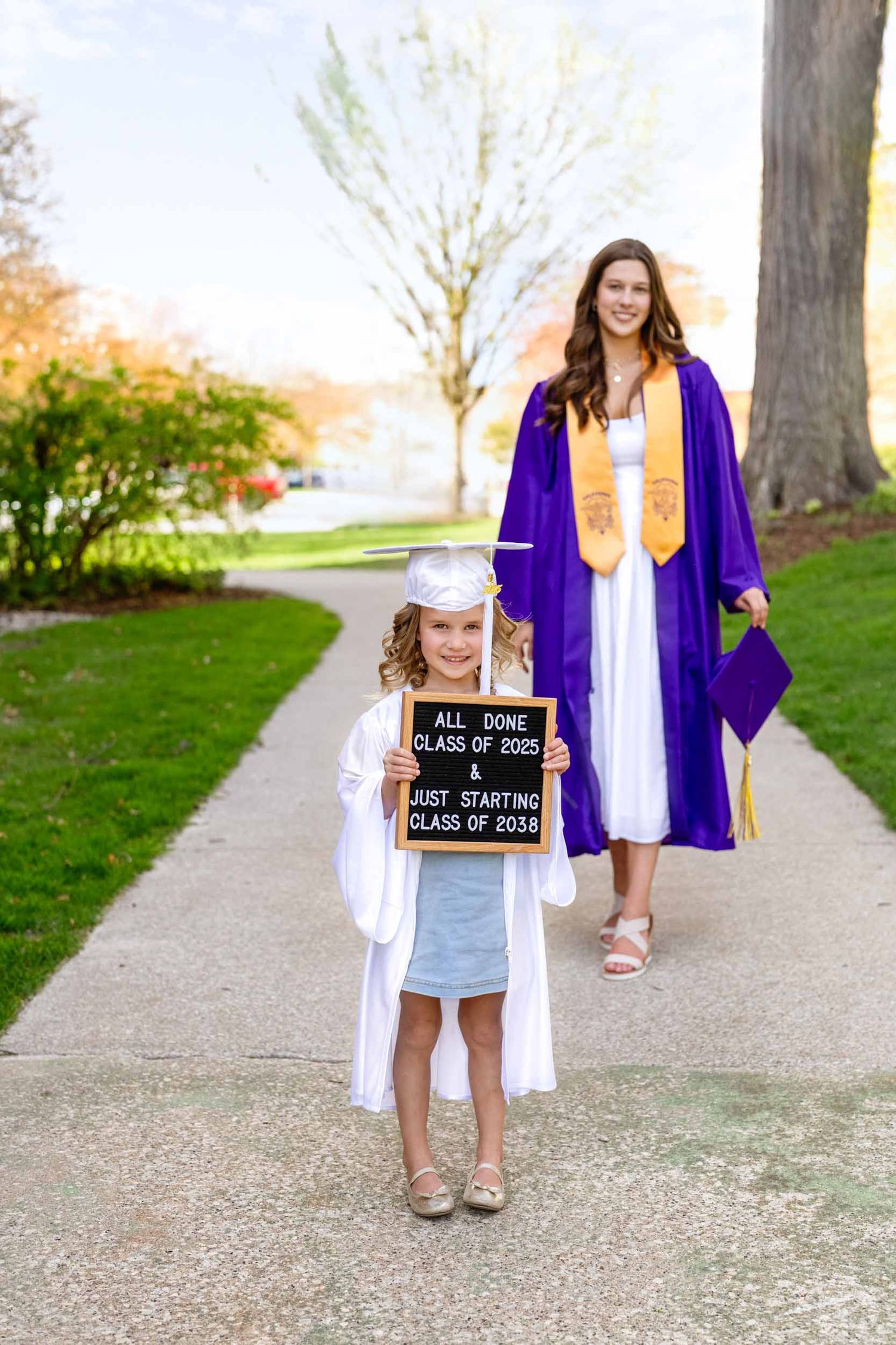 A graduation photo showing two graduates in white and purple robes posing on a walkway with trees in the background.