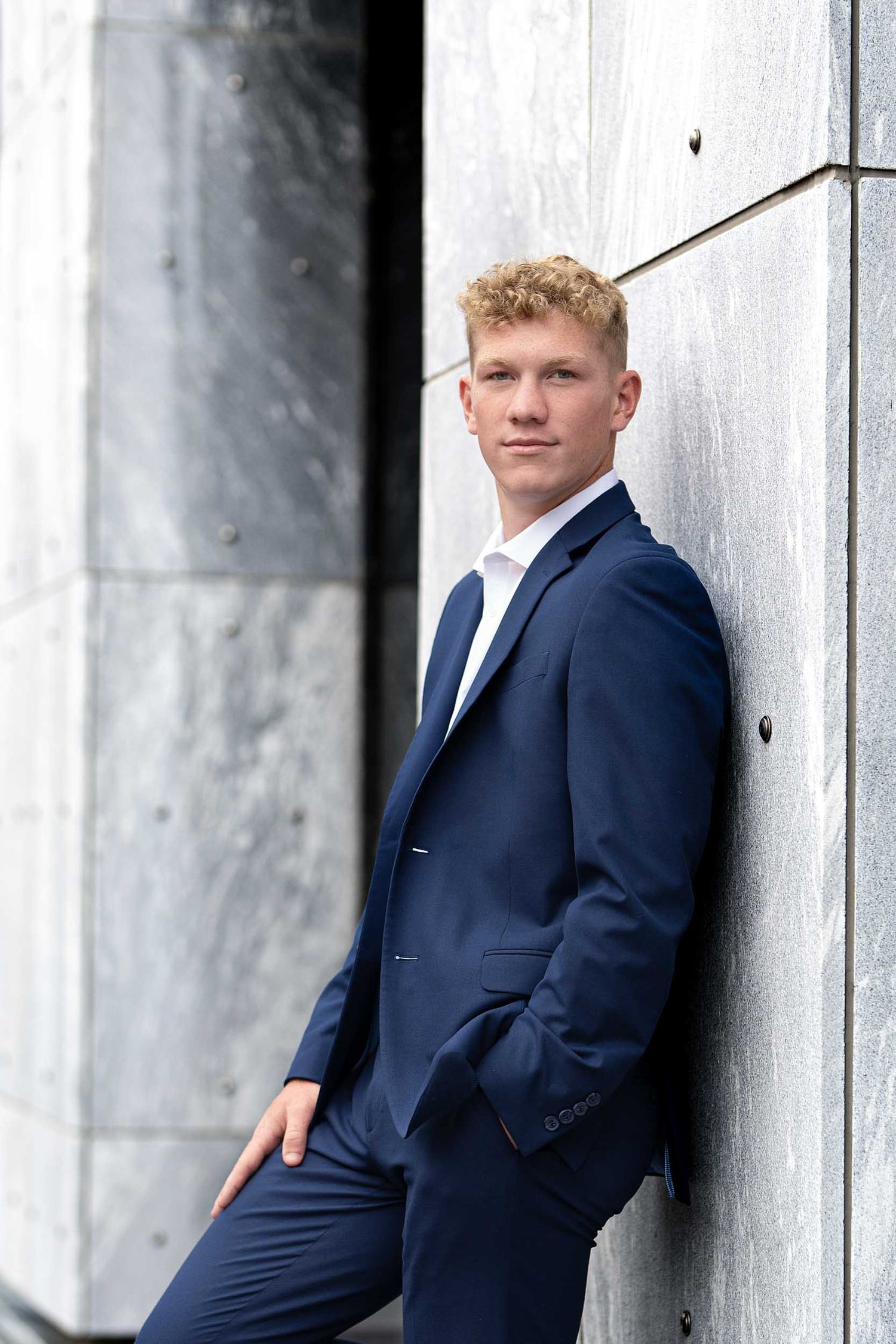 Young person in navy suit poses against white architectural columns in professional portrait series.