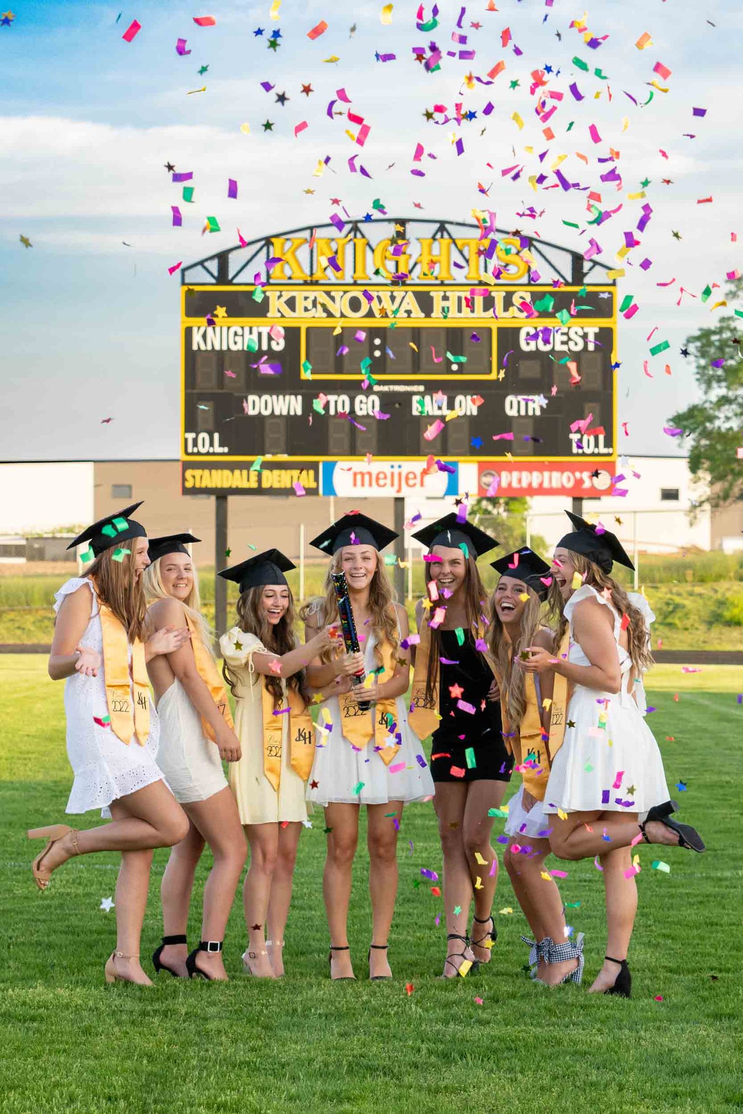 A group of graduates in white dresses celebrate with confetti under a graduation banner on a sunny field.