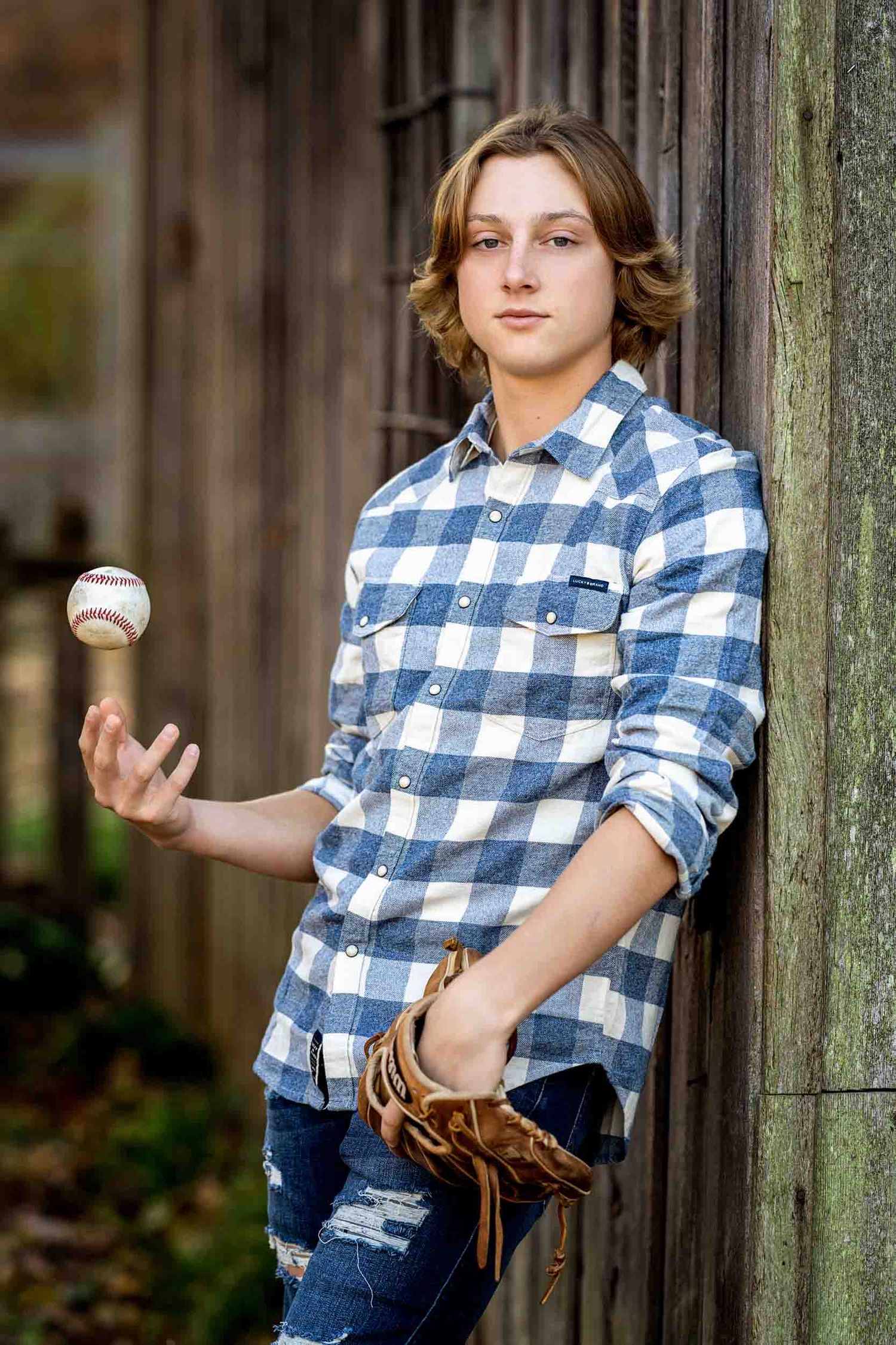 A youth in a blue plaid shirt and jeans practices baseball throwing poses against a rustic wooden fence background.