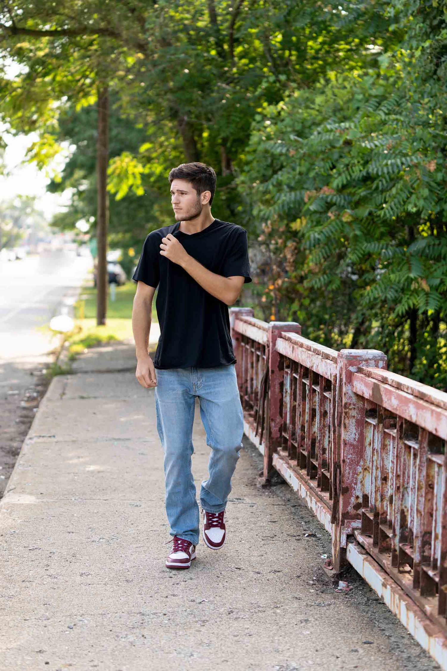 Person in black shirt and jeans walking along a rustic bridge railing in a park setting on a sunny day.
