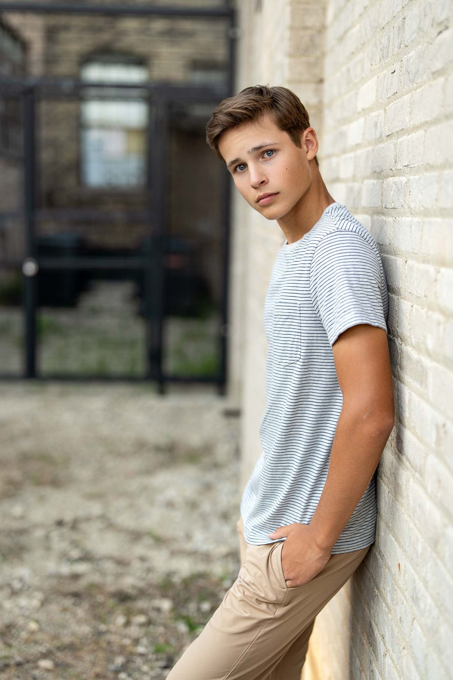 Young person in striped t-shirt and khaki pants leaning against brick wall in urban setting.
