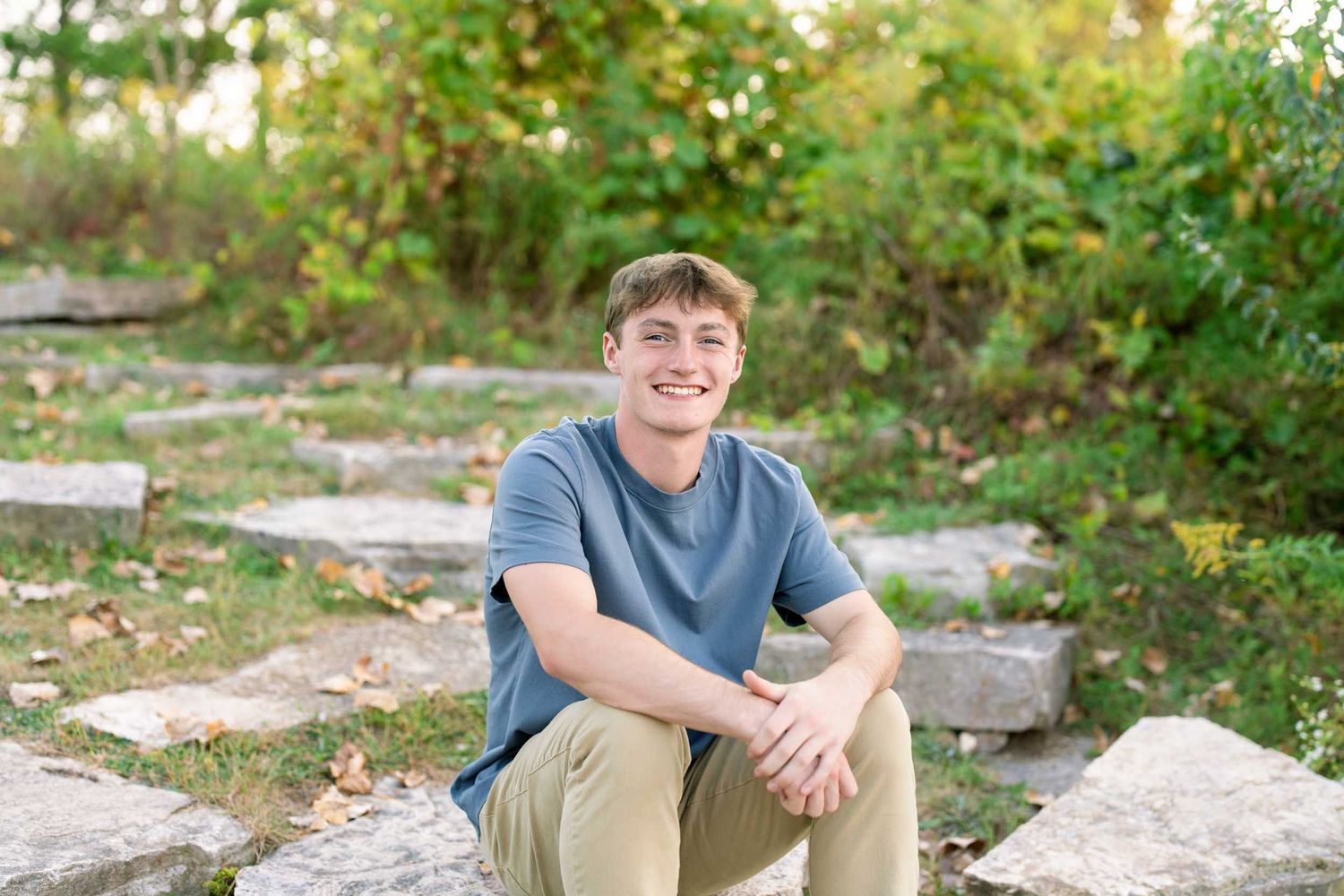 A person wearing a blue t-shirt sits casually on rocky terrain surrounded by greenery in an outdoor setting.