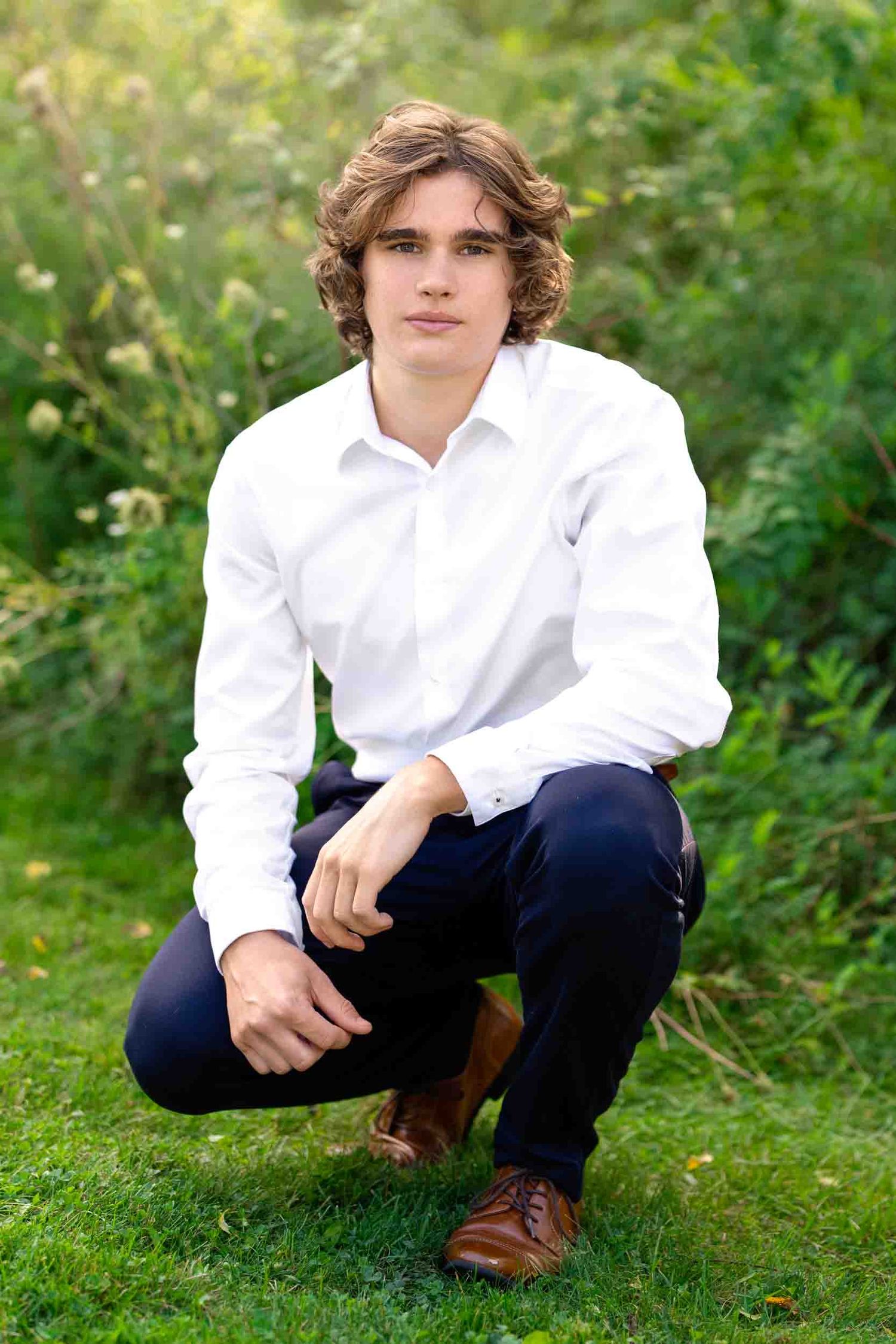 A person in a white button-down shirt and navy pants crouches in lush green grass wearing brown leather shoes.