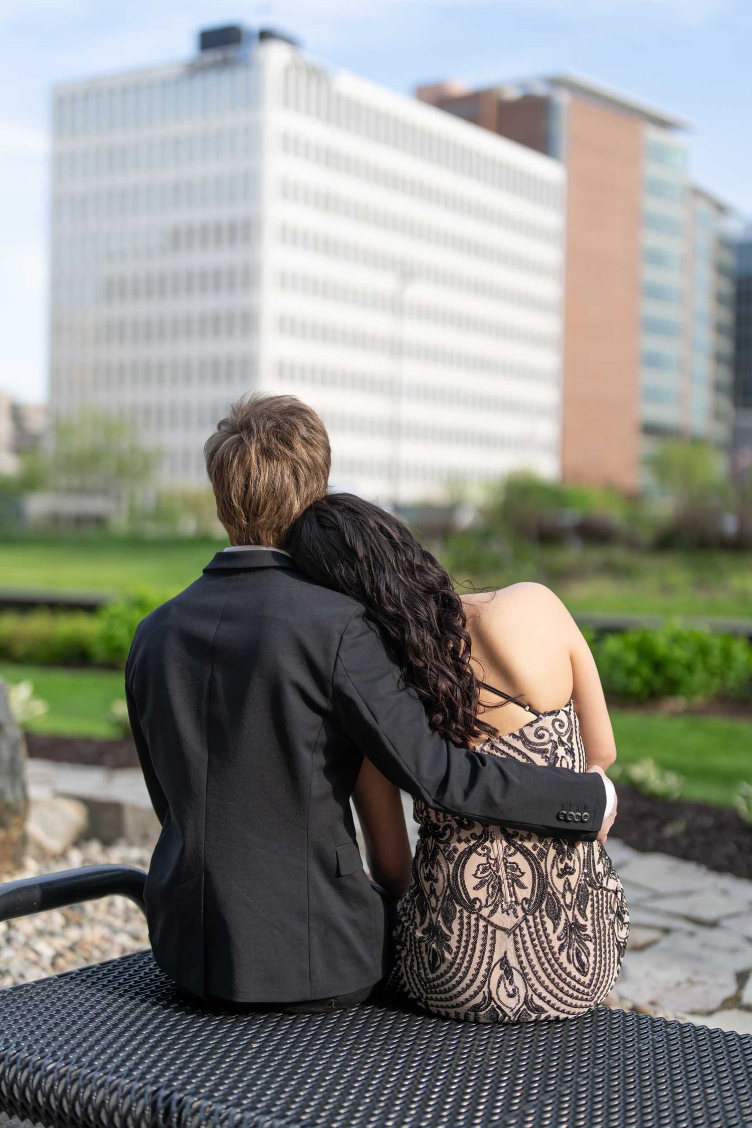 A couple sits together on a bench overlooking office buildings, with the person wearing a patterned dress leaning on their partner.