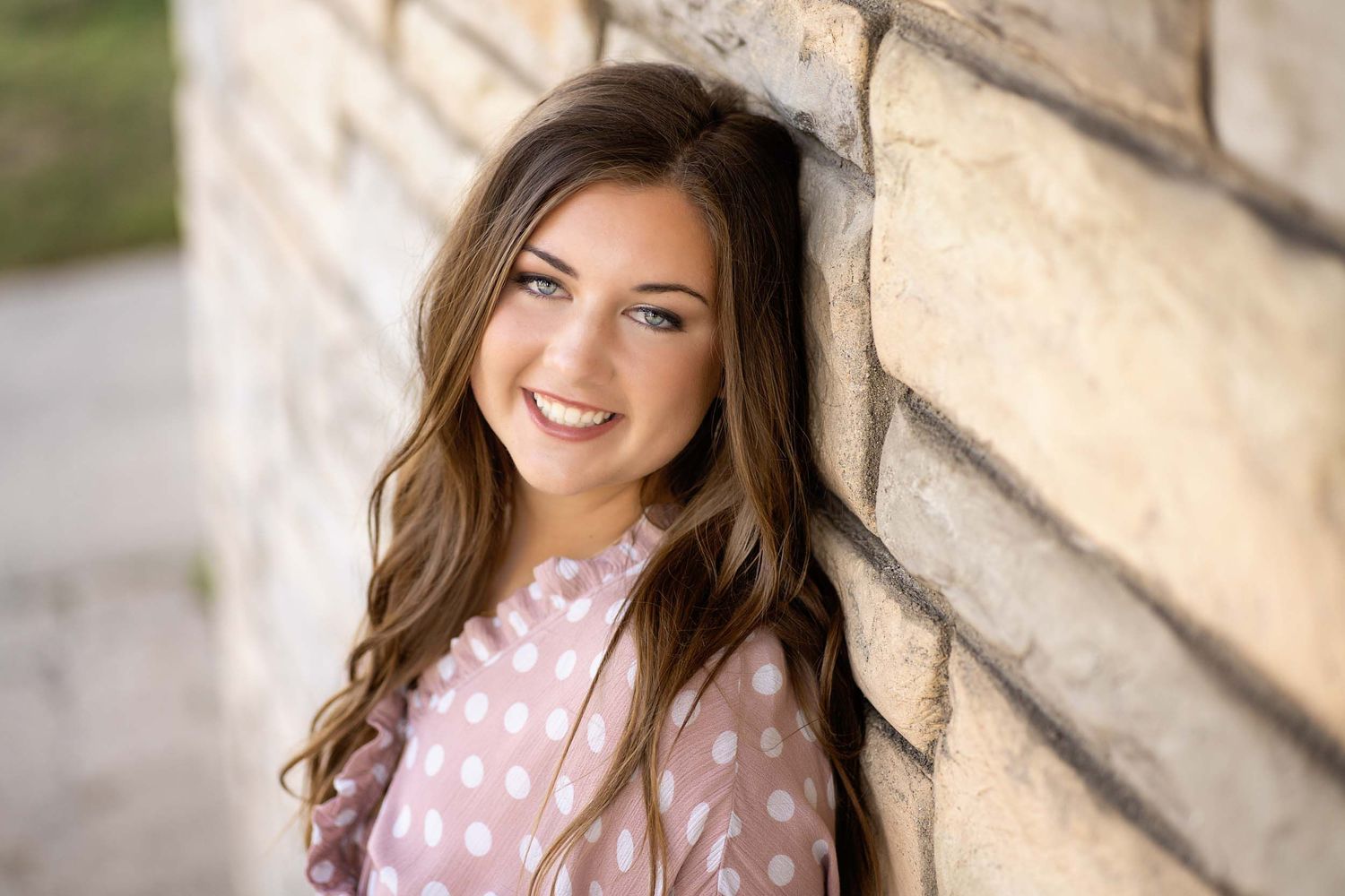 A person in a pink polka dot dress smiles while posing against a light-colored stone wall.