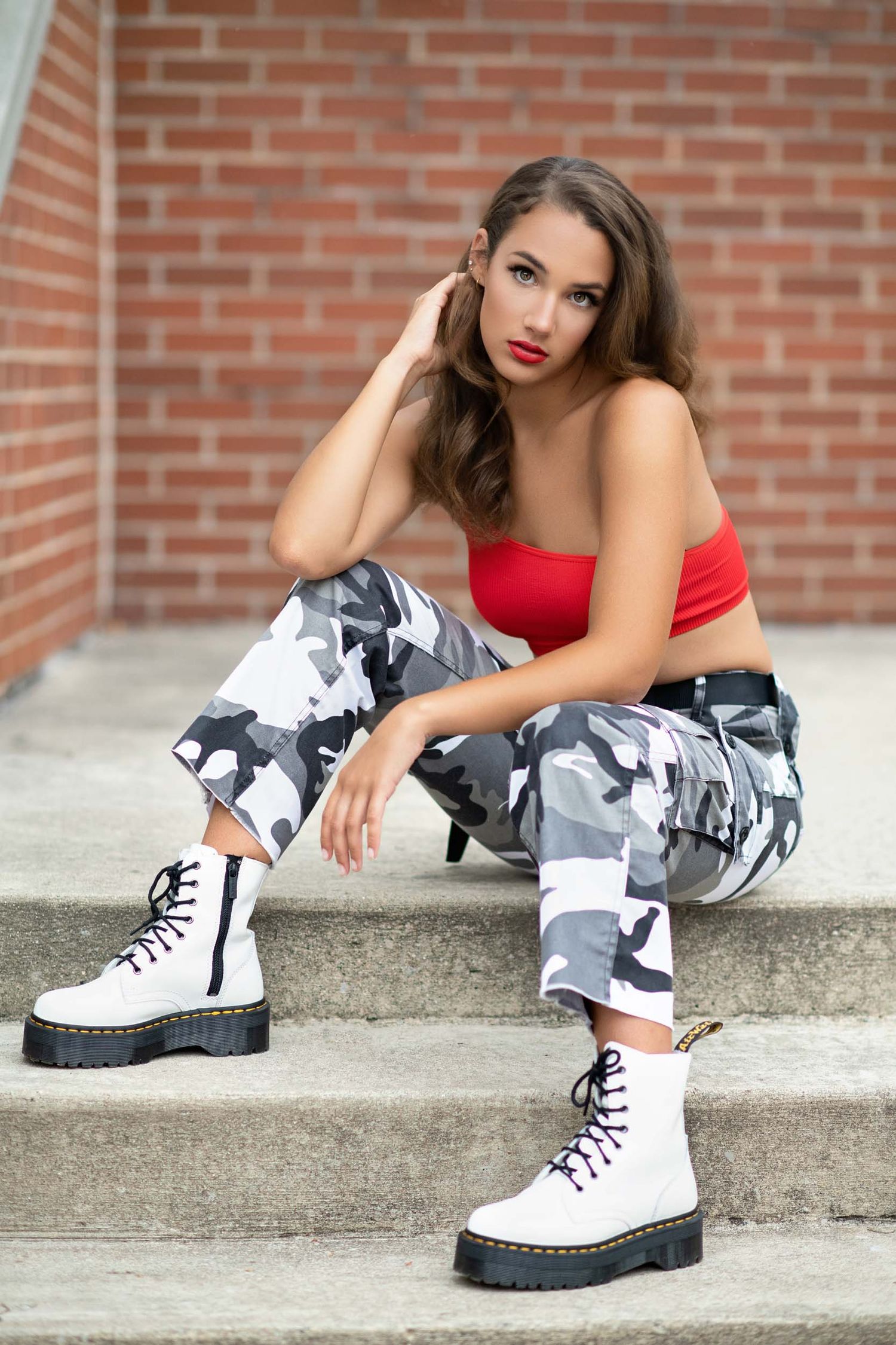 Fashion model wearing red top and camo pants poses with white combat boots against brick wall.