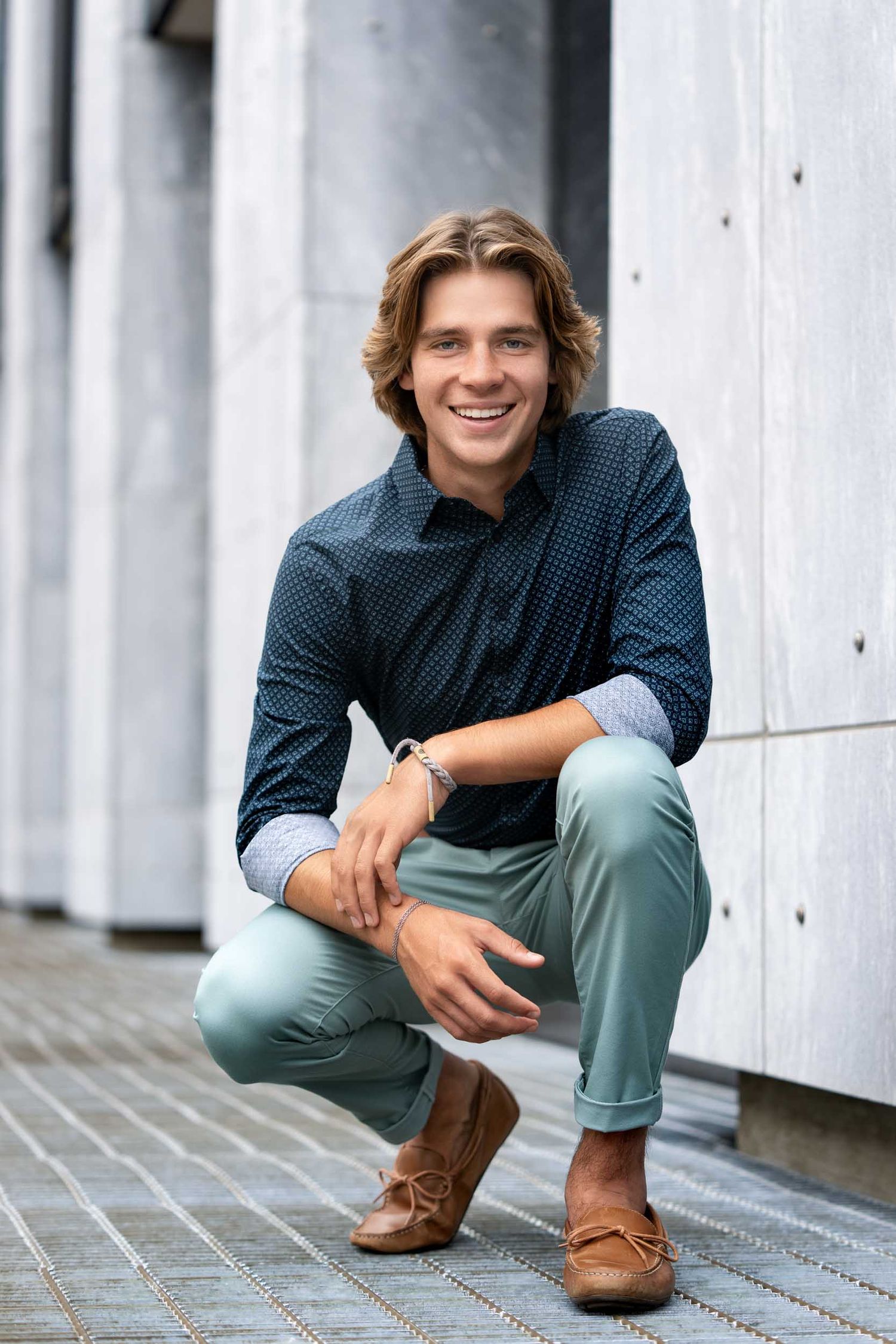 A person in a blue polka dot shirt and mint green pants crouches casually against a white column background.
