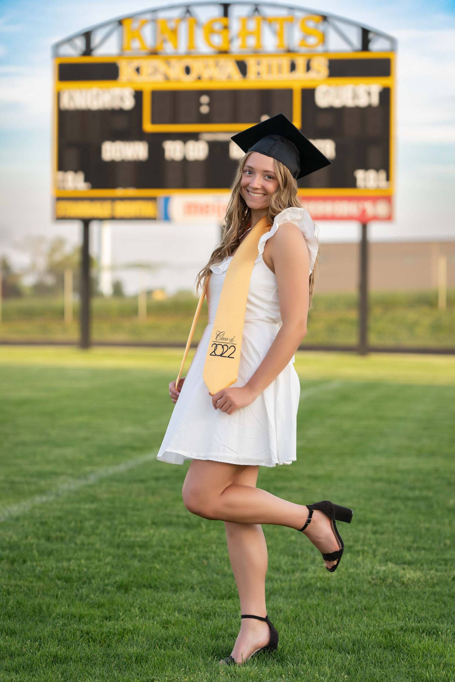 Graduate in white dress and black graduation cap poses playfully on football field with scoreboard in background.