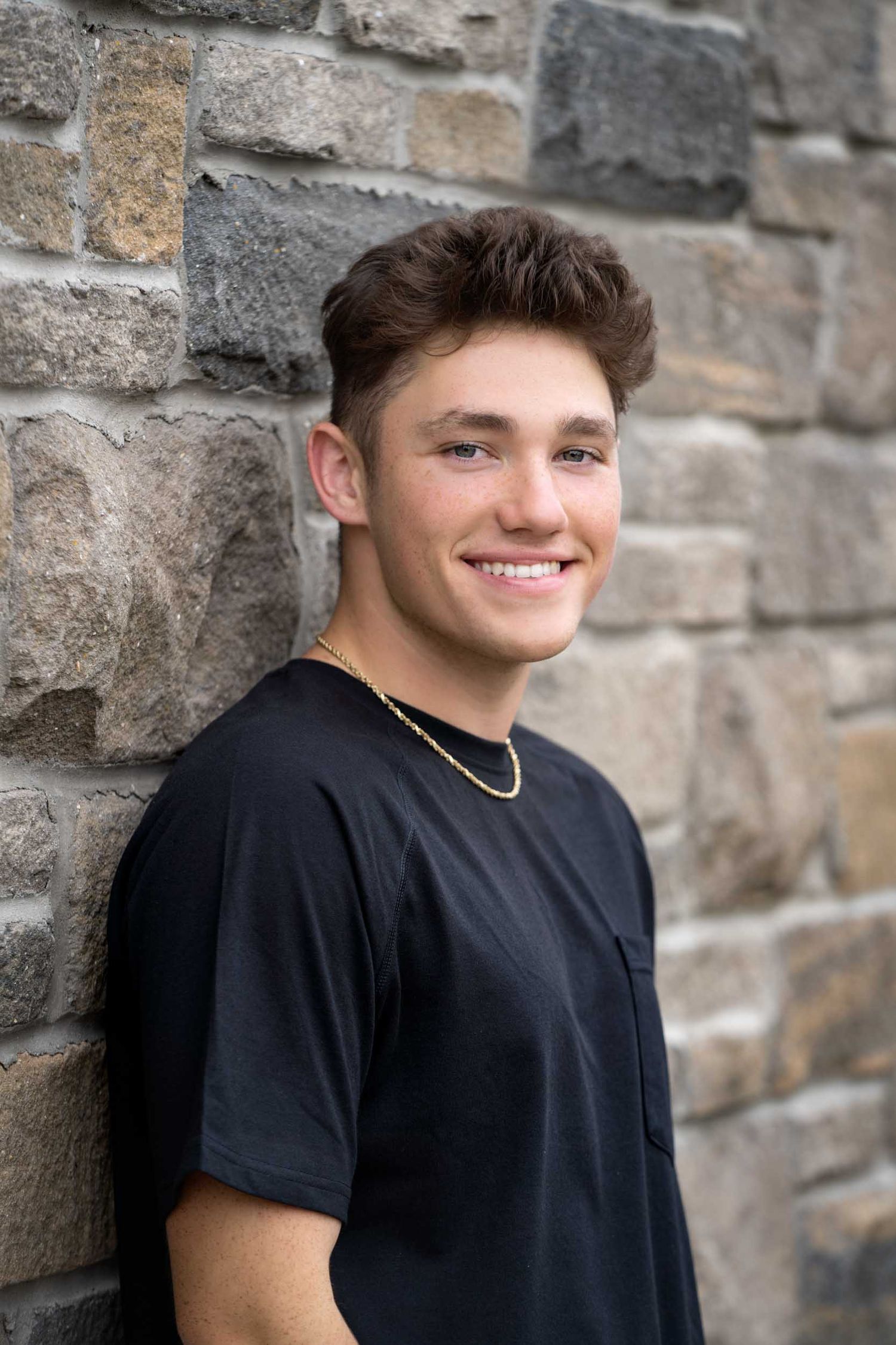 A young person in a black t-shirt and gold chain smiles while leaning against a stone wall outdoors.