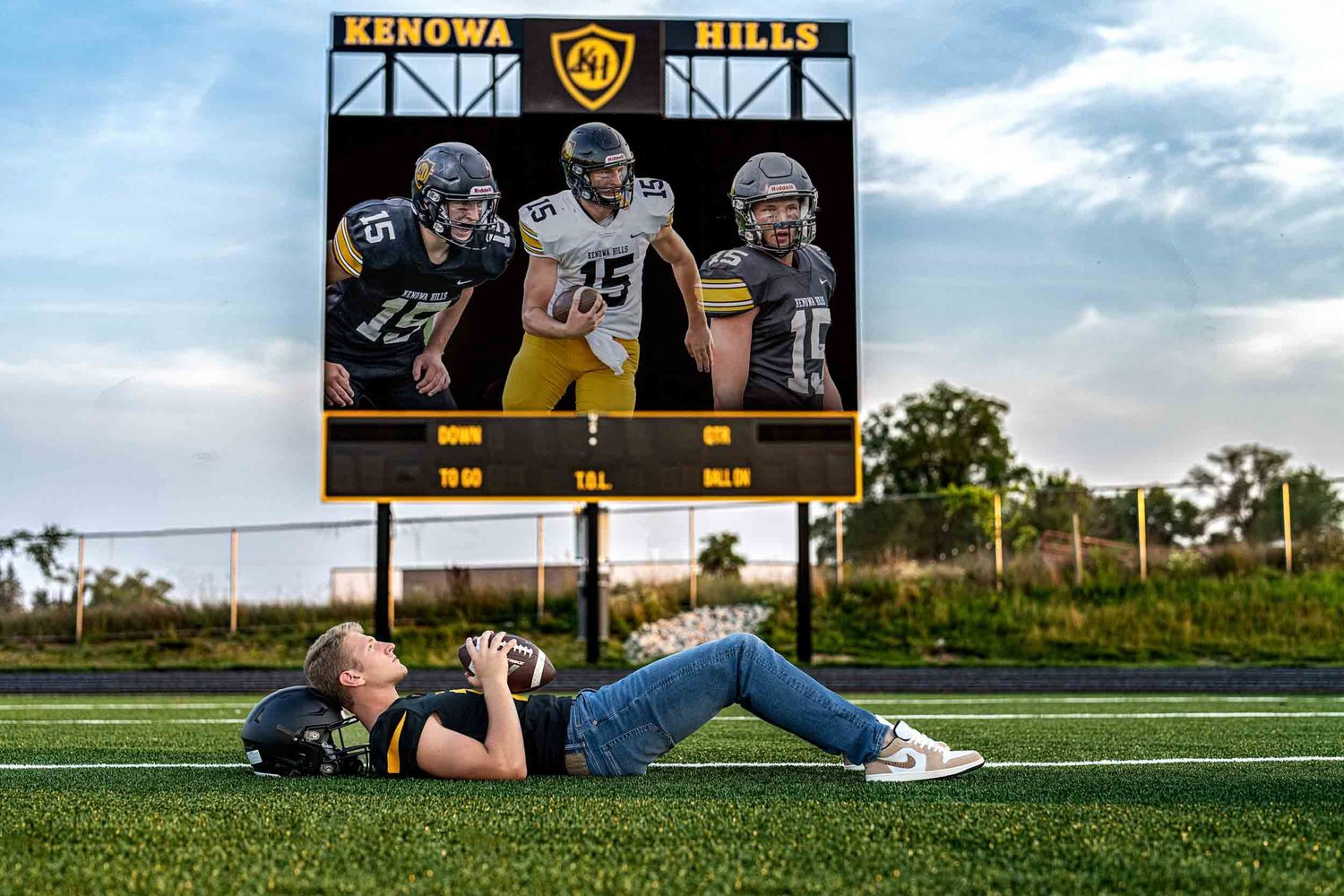 A photographer lies on a football field capturing photos with a digital scoreboard display showing athletes in the background.