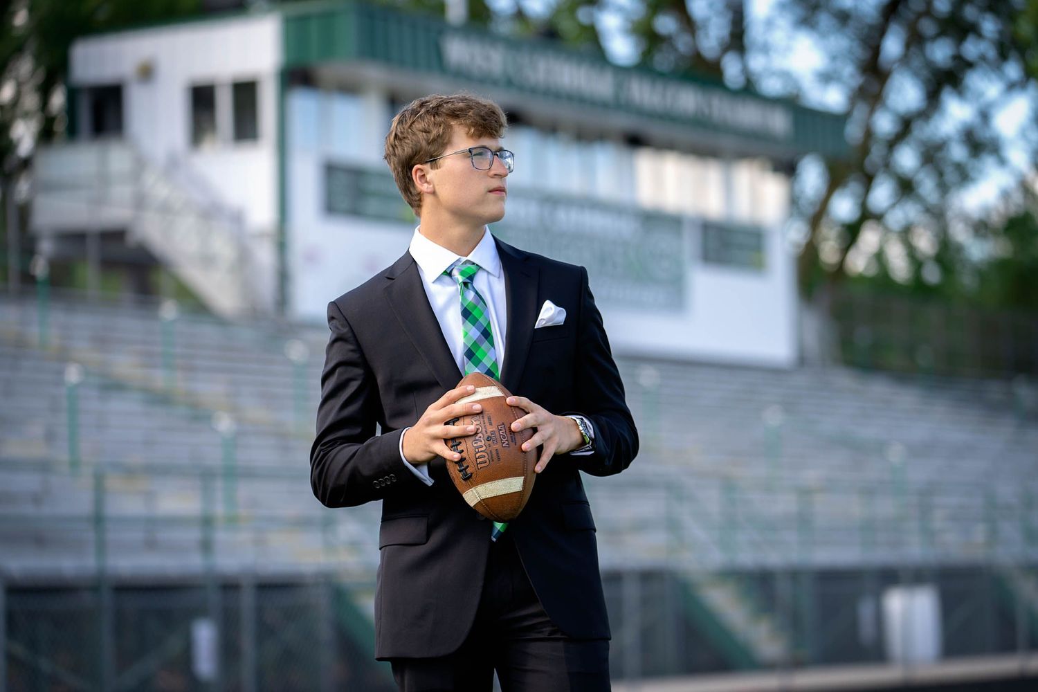 High school senior in formal attire poses with football on bleachers for senior portraits.