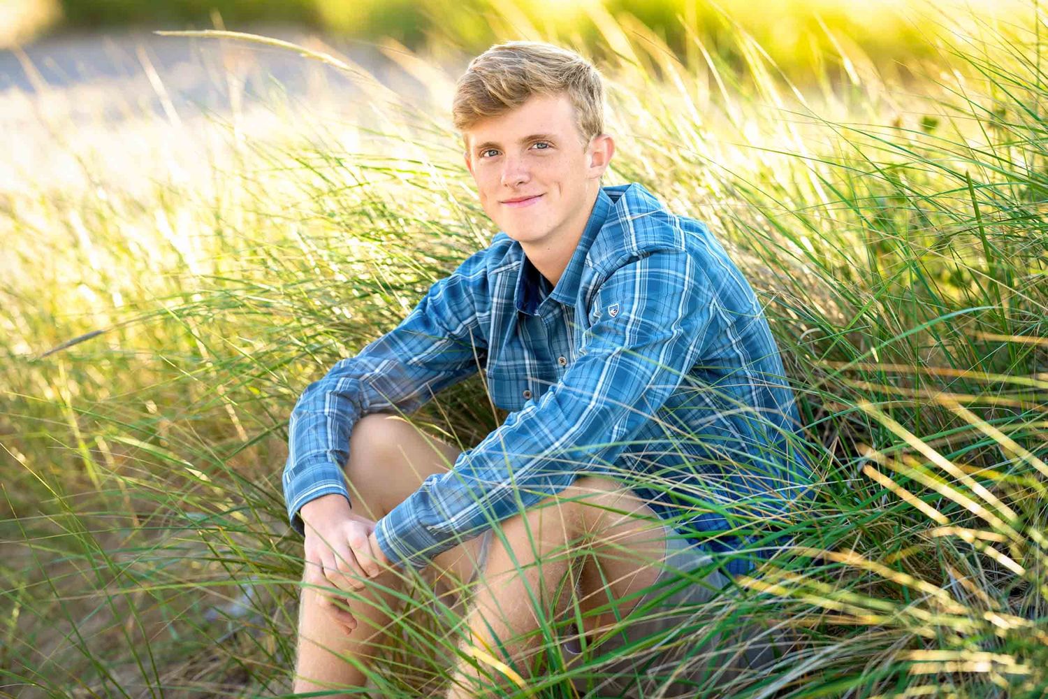 A person in a blue plaid shirt sits in tall grass during golden hour sunset lighting.
