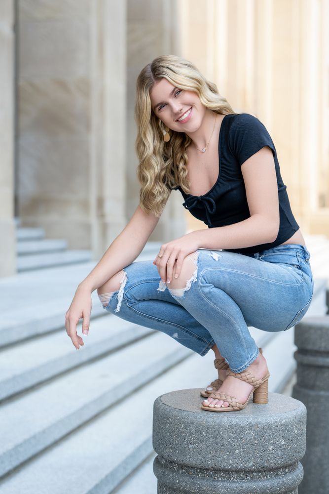 Person in casual denim and black top poses on concrete pillar with elegant architectural backdrop.
