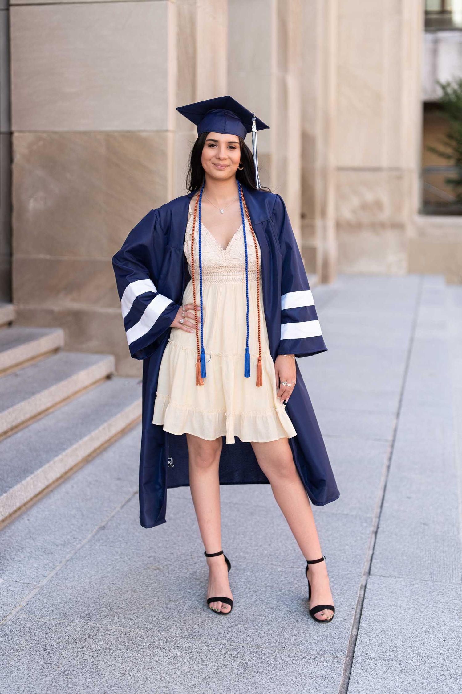 Graduate in navy cap and gown poses on concrete steps in flowing beige dress.
