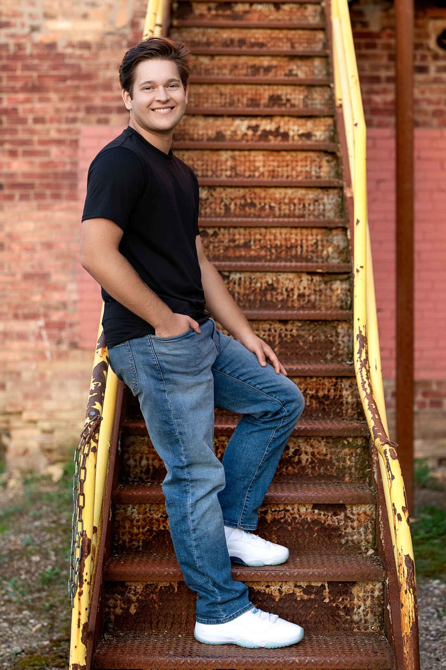 A person in a black t-shirt and blue jeans poses casually on rusty metal stairs against a brick wall.