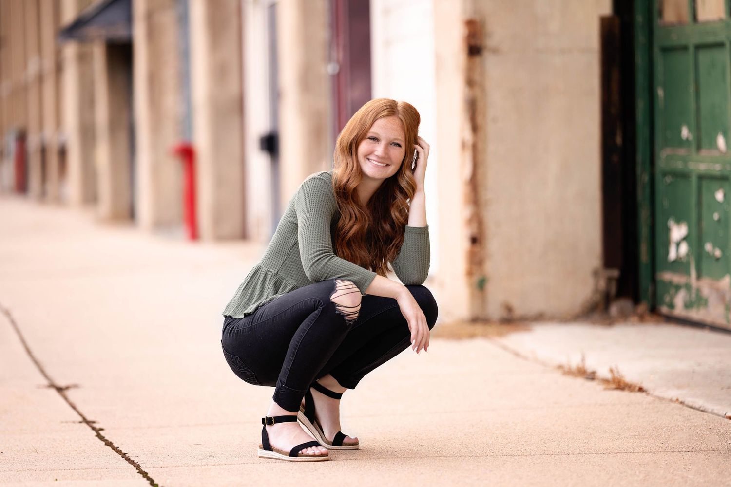 A person in casual attire poses on a city sidewalk with stone walls and green doors in the background.