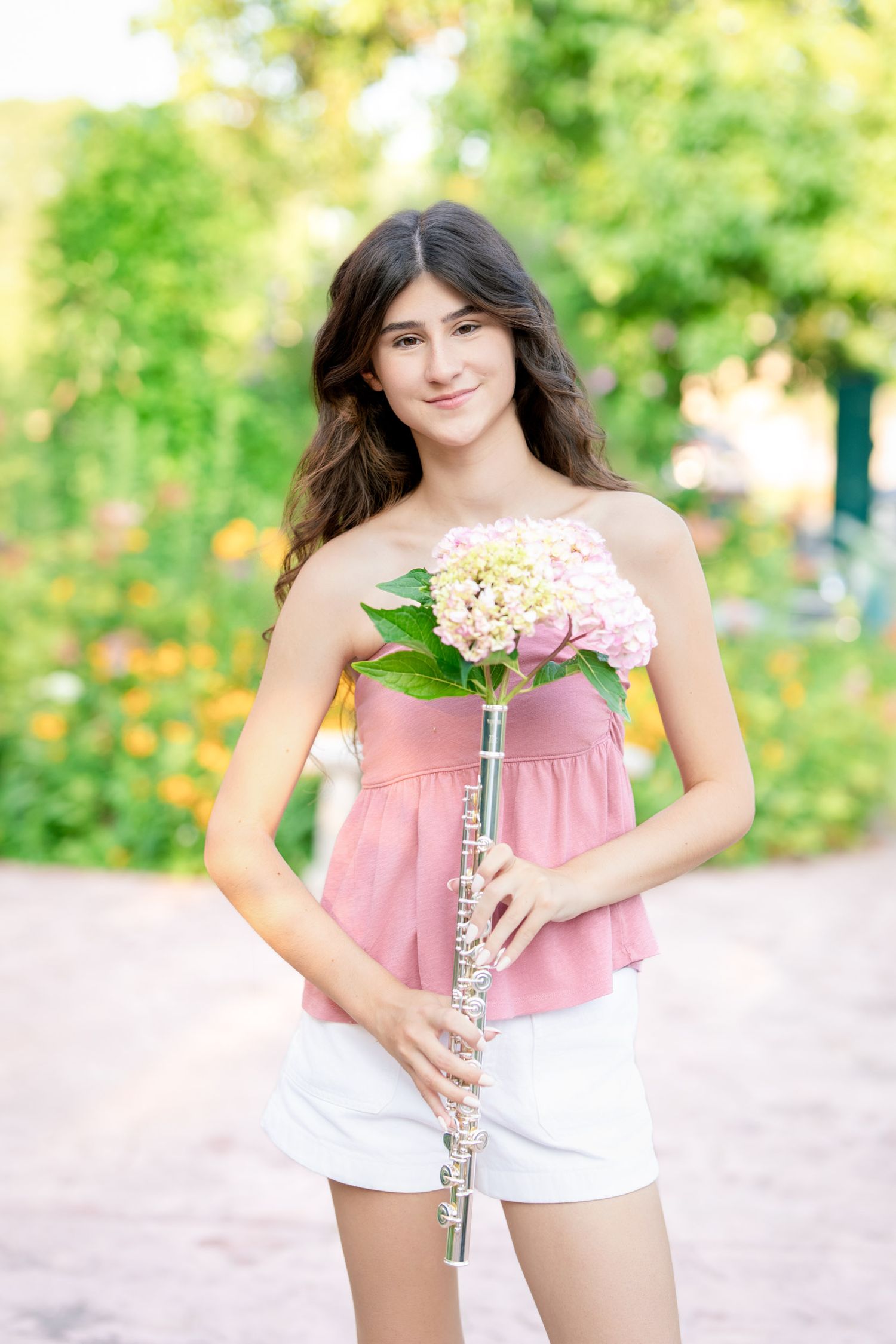 A musician in pink holding a flute poses in a garden with flowers.