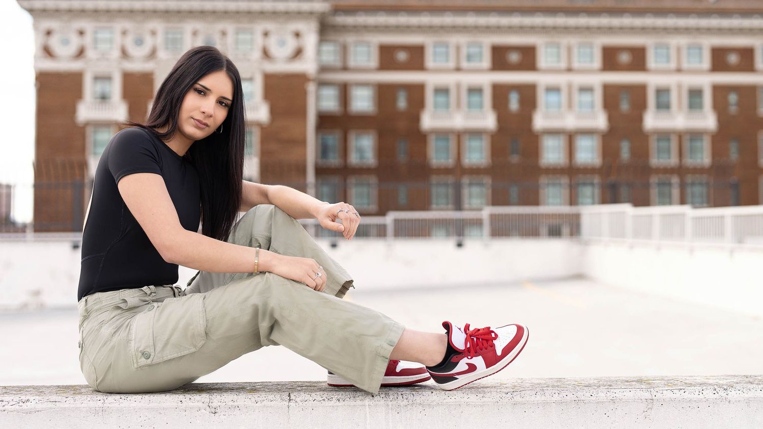 Young woman in black tee, cargo pants, and red Nike sneakers sitting outdoors near a brick building.