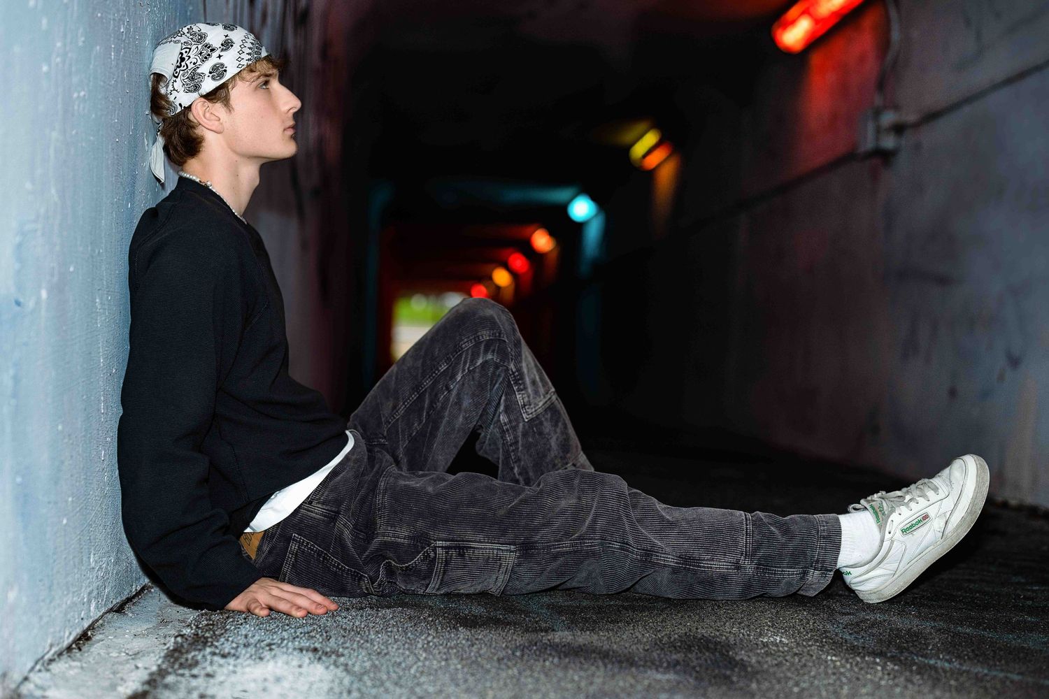 Young person in black outfit and bandana sits against tunnel wall with colorful lights in background.