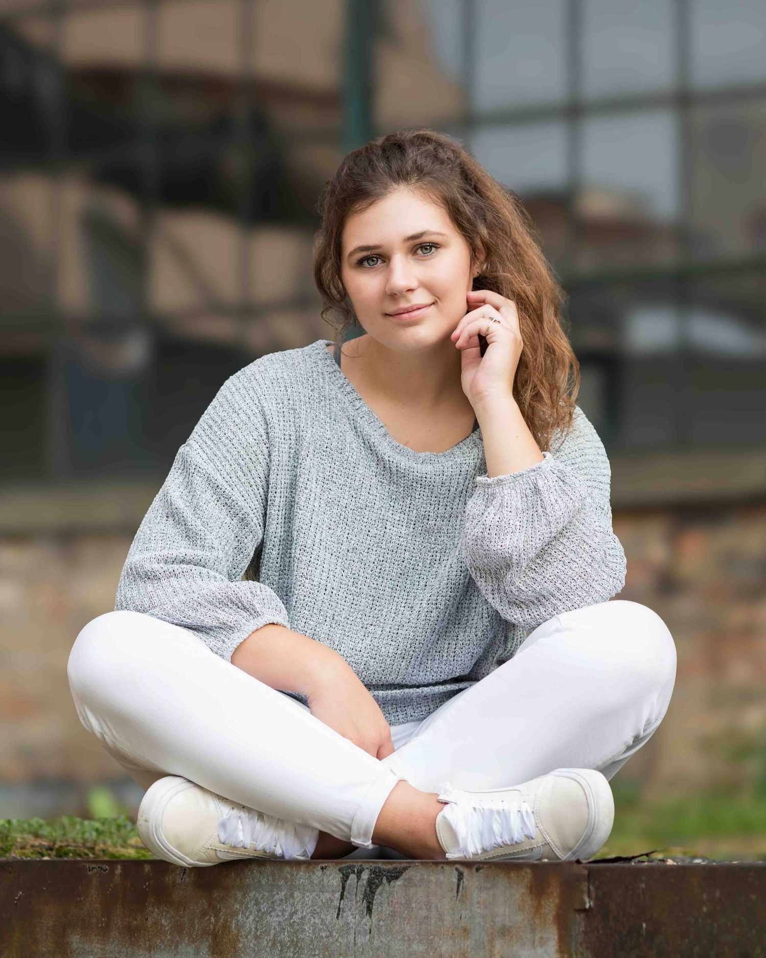 Young woman in gray sweater and white jeans sitting cross-legged outdoors, smiling with hand near face.