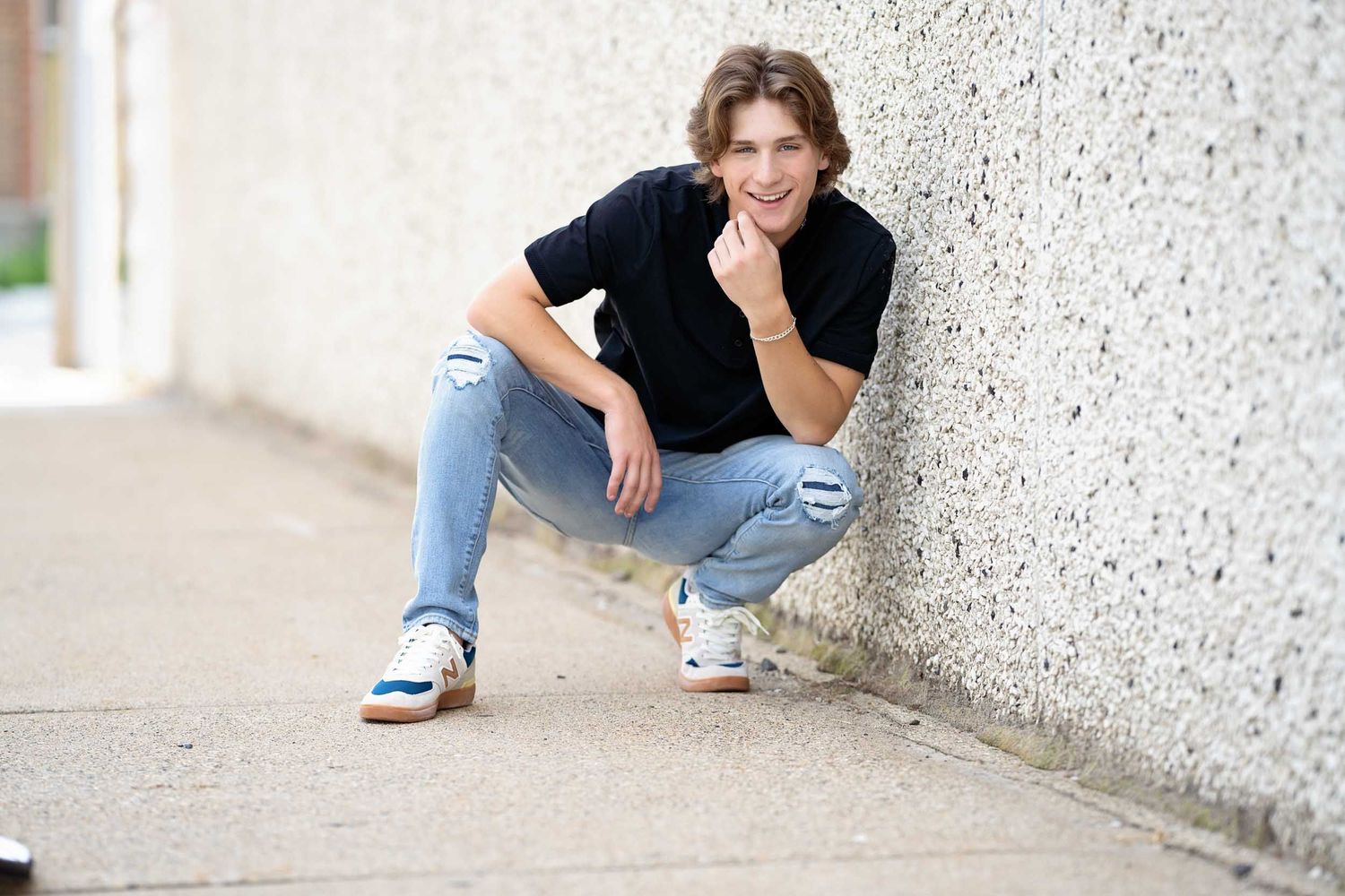 A casual figure in a black t-shirt and ripped jeans crouches against a textured white wall outdoors.