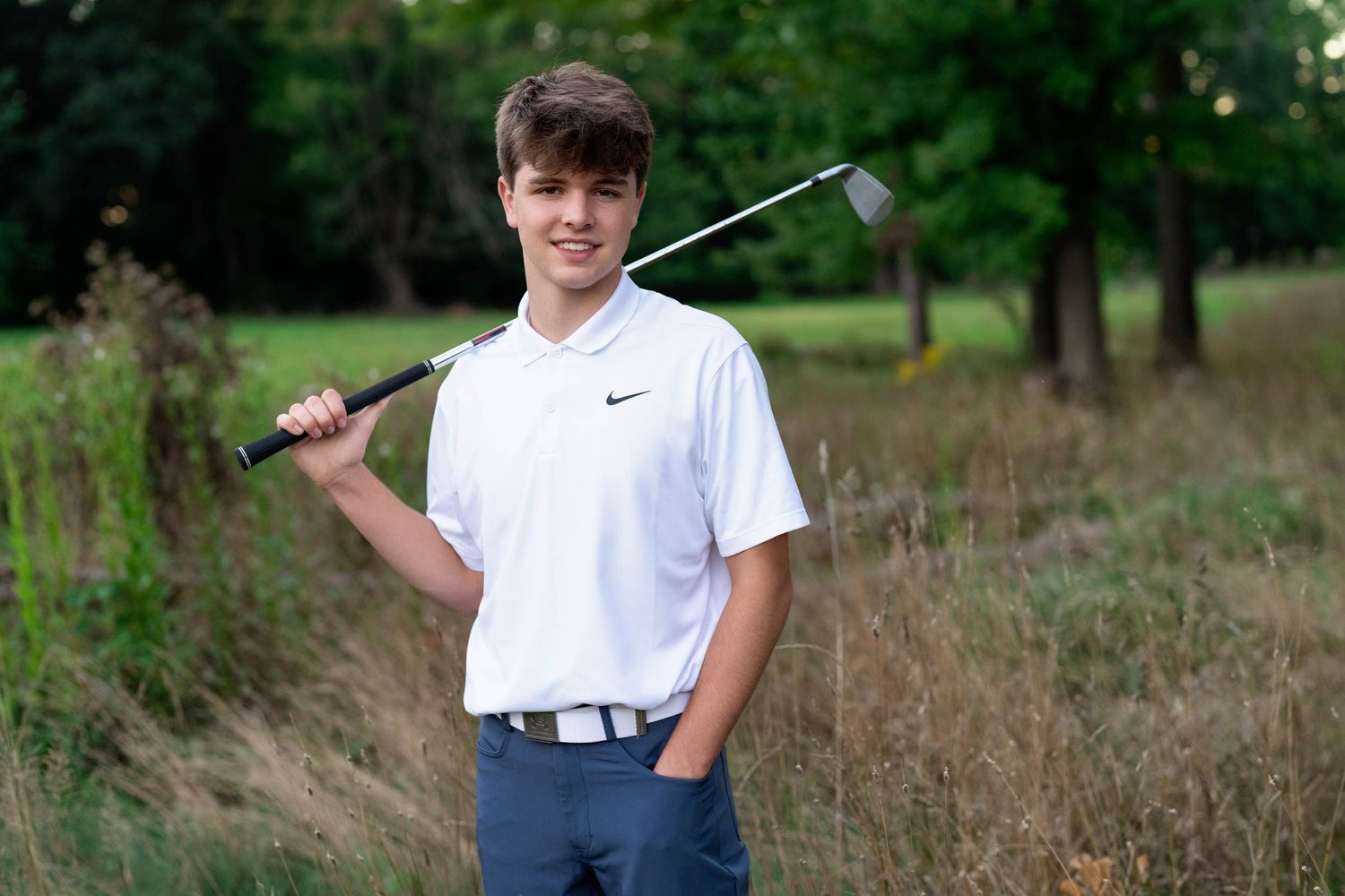 A golfer poses with a club on a lush golf course surrounded by trees and natural scenery.