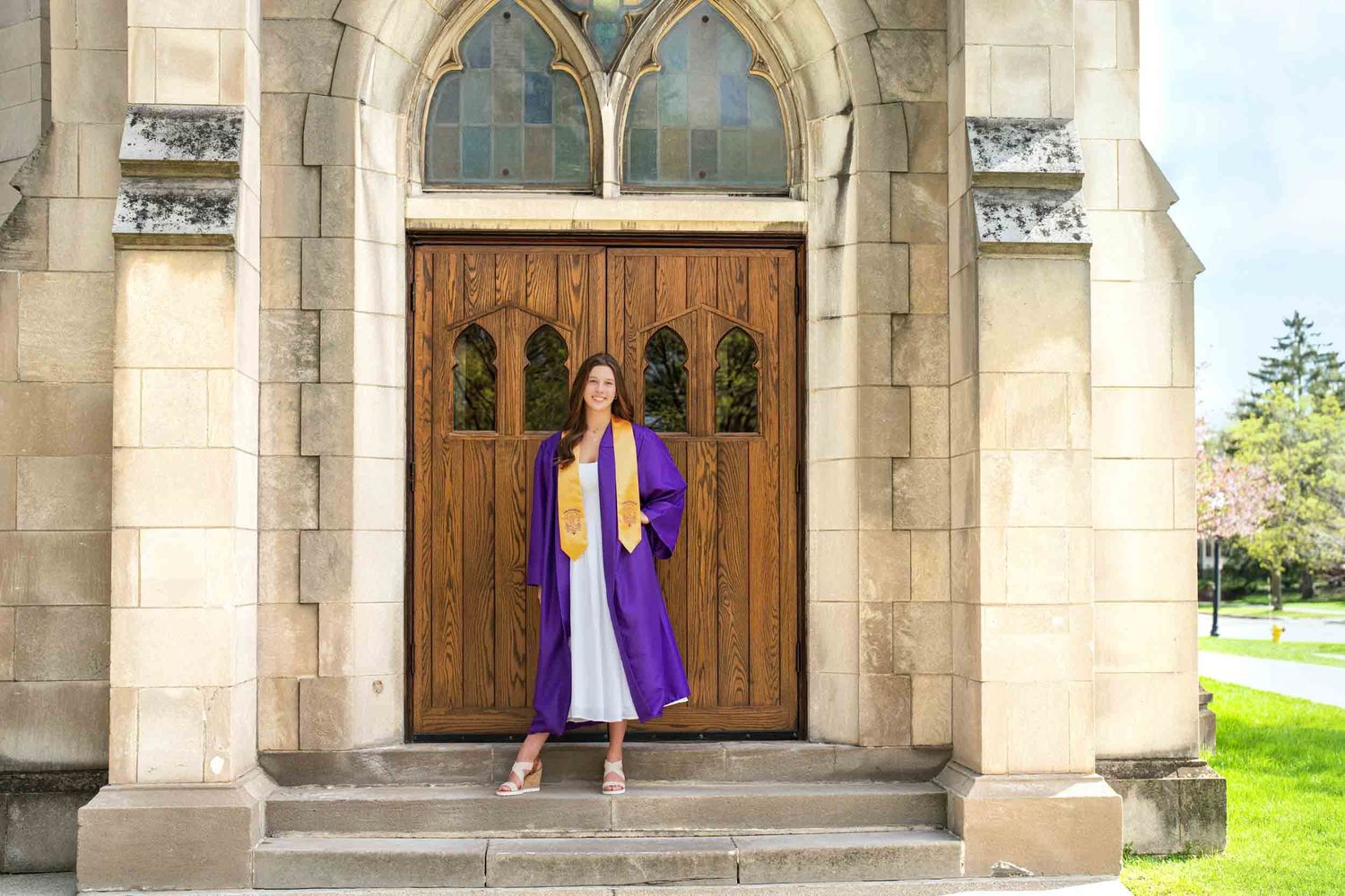 A graduate in purple and gold regalia stands in front of ornate wooden church doors with gothic stone archways.