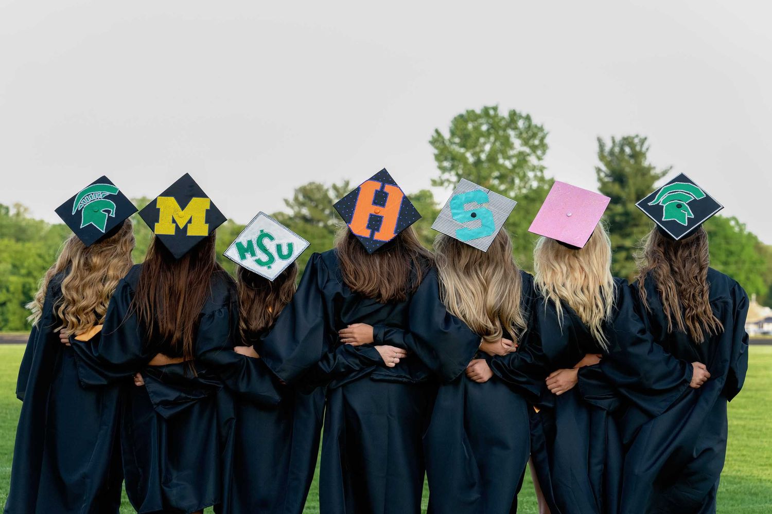 A row of graduates in black caps and gowns photographed from behind during an outdoor graduation ceremony.