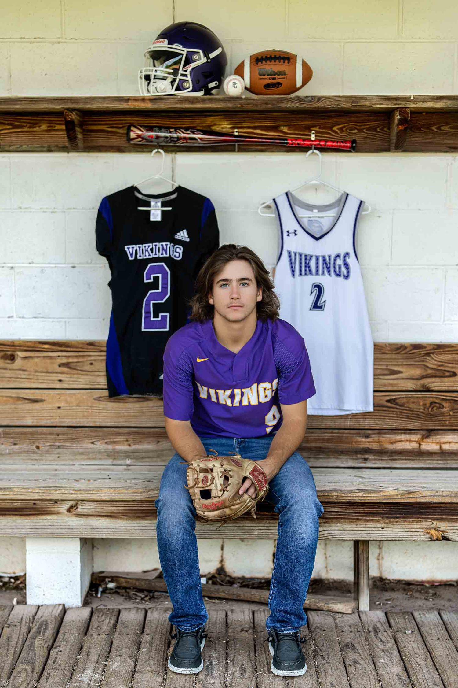 High school football player sits on wooden bleachers with Vikings jerseys displayed behind them.