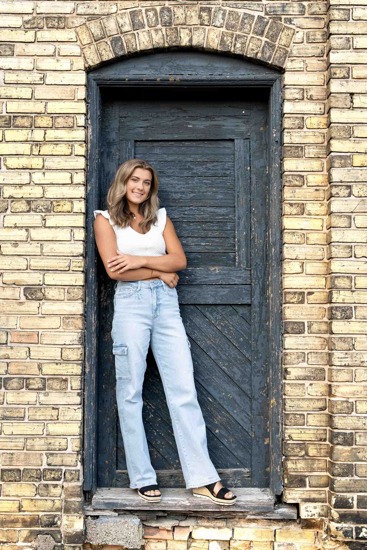 Person in light jeans and white top sits in rustic brick window frames with vintage blue doors.