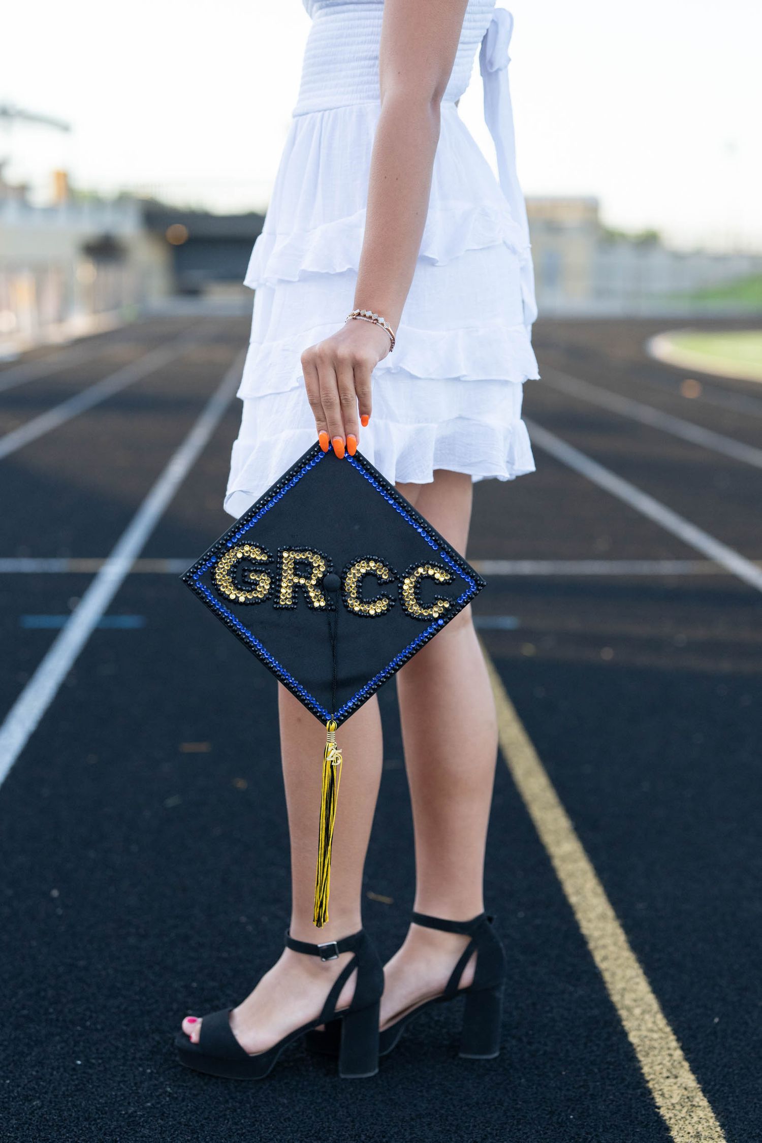 A graduation cap decorated with gold letters 'GRCC' sits against a black asphalt background with painted white lines.