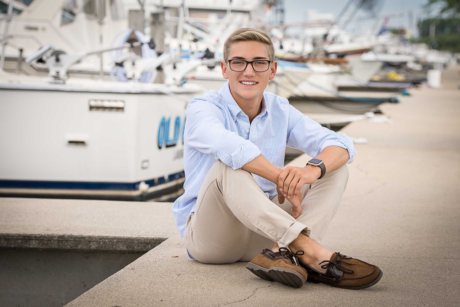 A person wearing glasses and a light blue button-down shirt sits casually on a dock near boats at a marina.