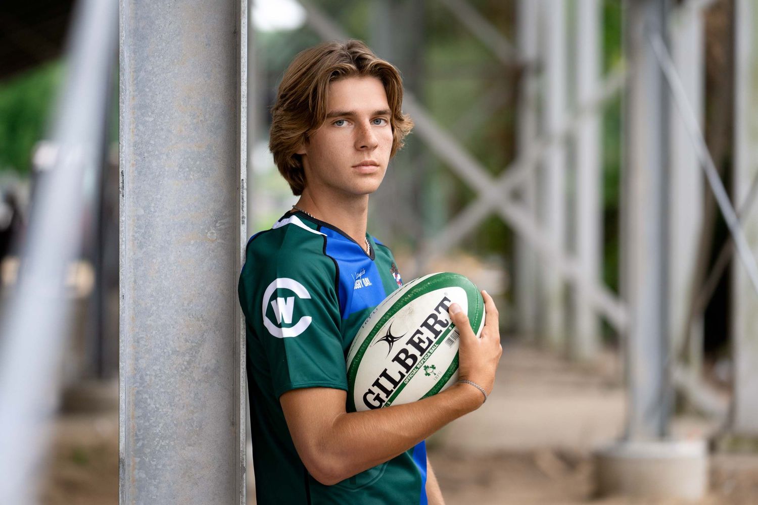 A rugby player in a green jersey holds a Gilbert rugby ball while standing against a blurred outdoor background.