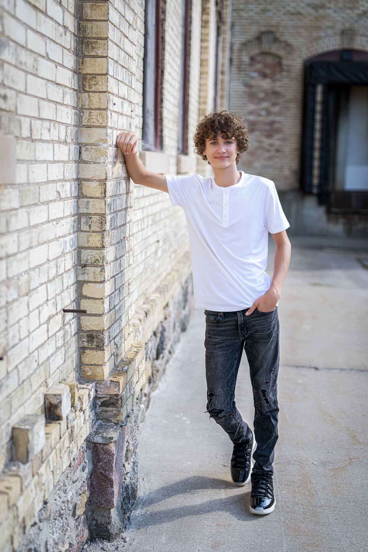 Young person in white t-shirt and dark jeans leans against brick wall in urban setting.