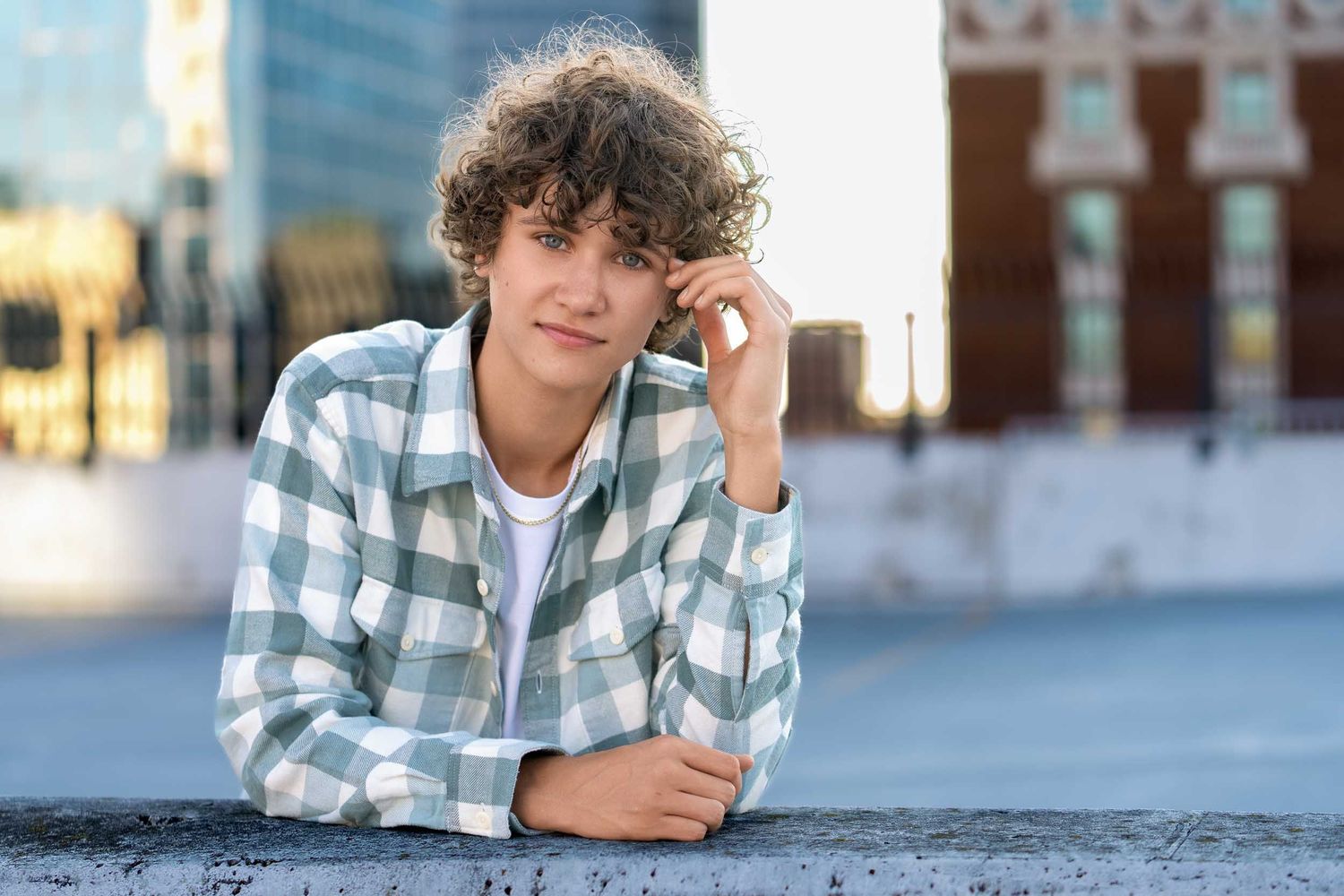 Young person in plaid shirt and curly hair leaning on concrete surface in urban setting during daytime.