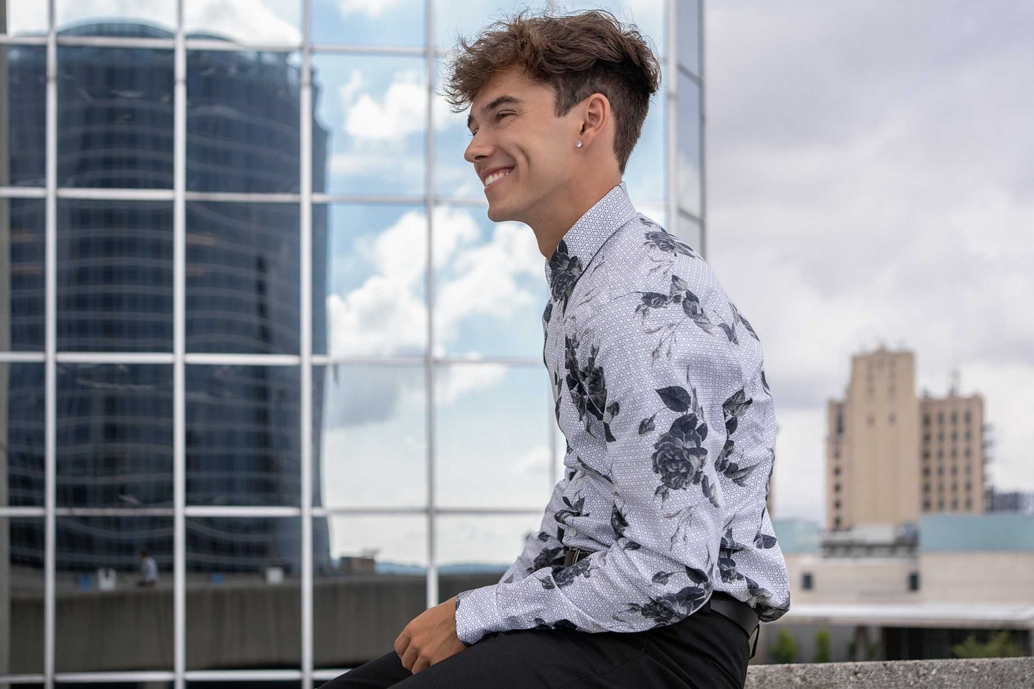 Professional in a white floral print dress shirt stands near reflective office building windows on a sunny day.