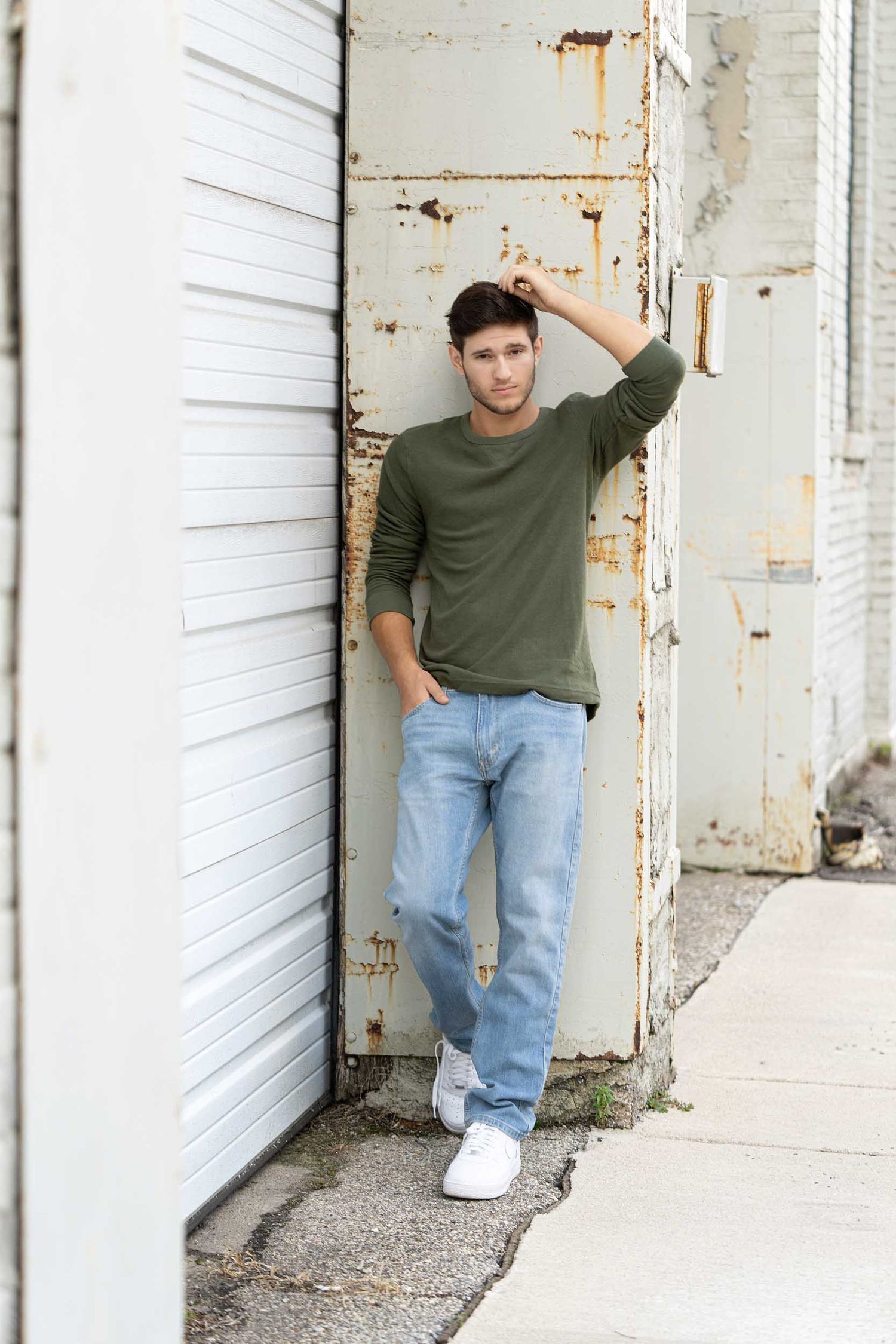 A person in a green sweater and light jeans poses casually against a white storage unit door.
