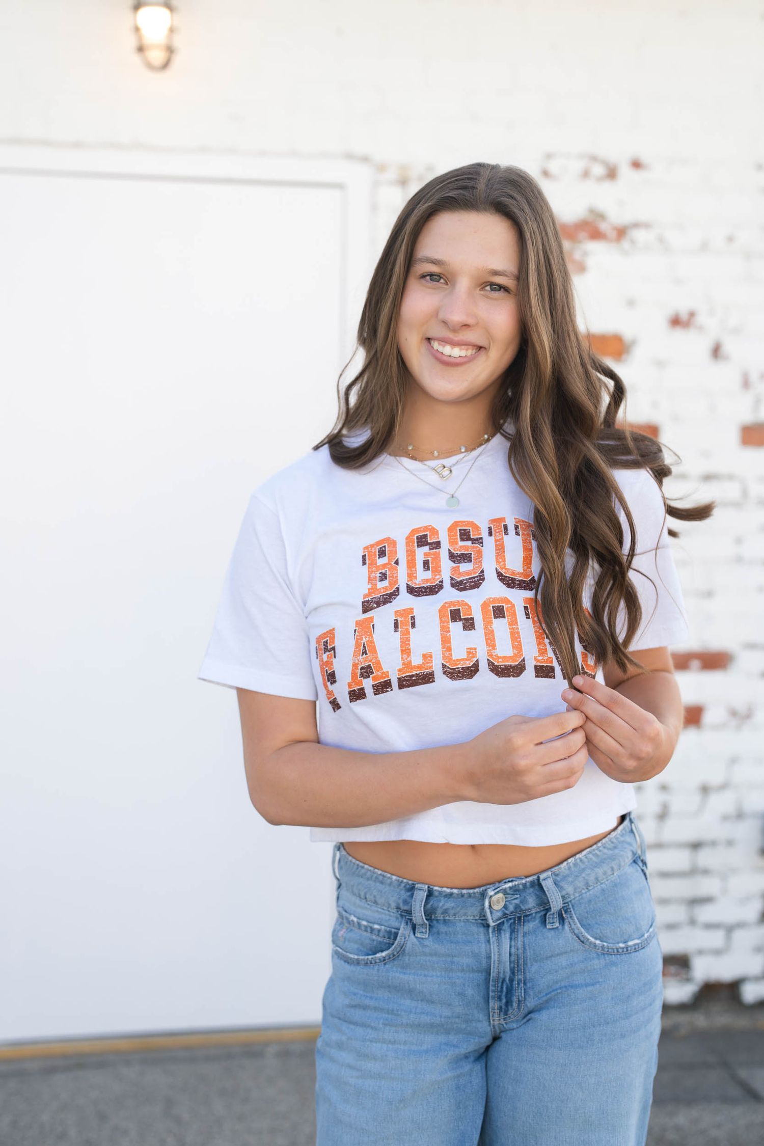 Student wearing a white BGSU Falcons cropped t-shirt and light wash jeans poses against white brick wall.