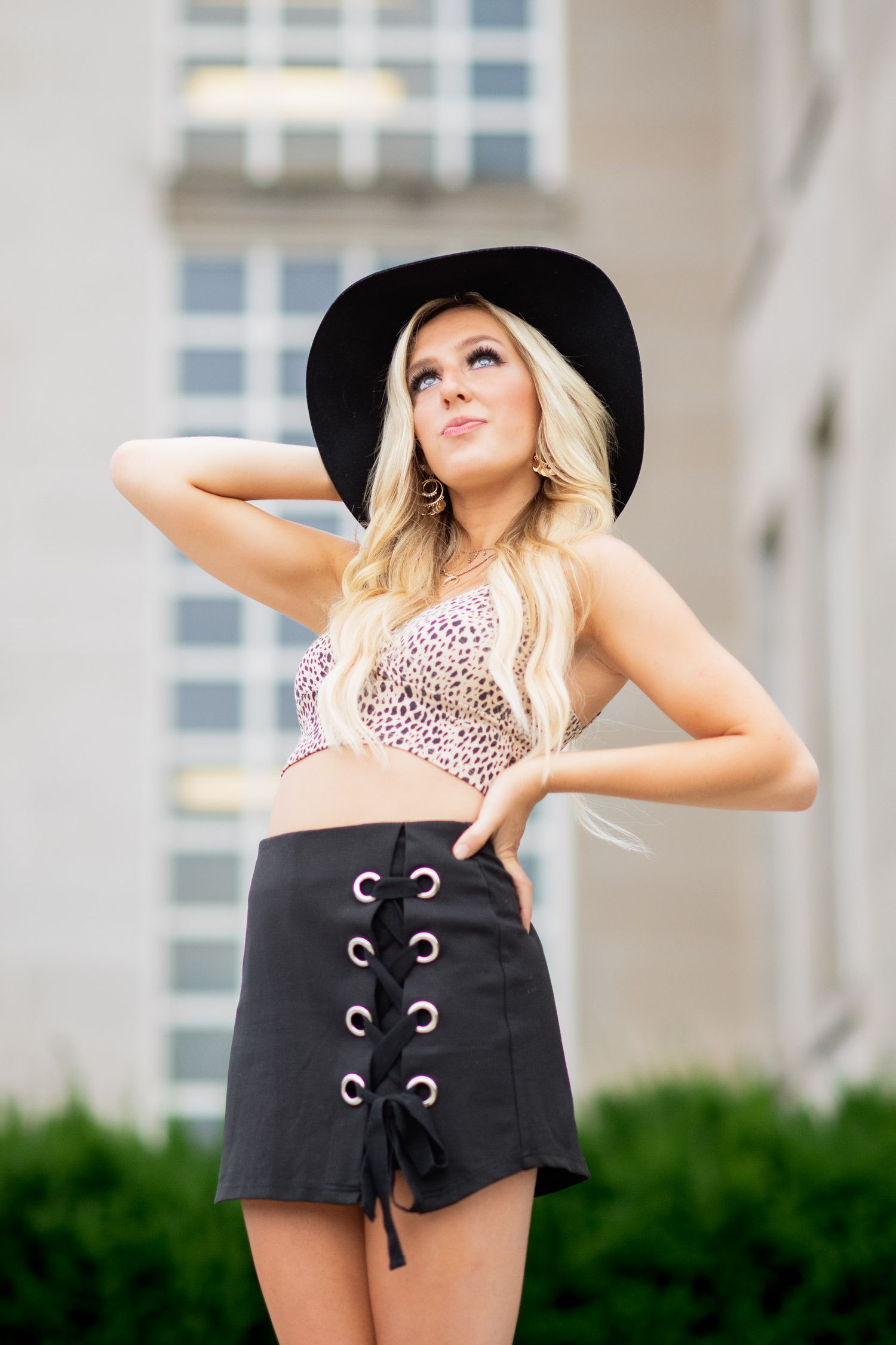A woman in a pink leopard print top and black shorts poses outdoors against a building with greenery.