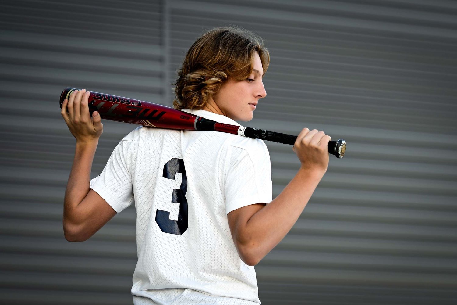 Baseball player in white jersey with number 3 holds a bat across shoulders against gray wall background.