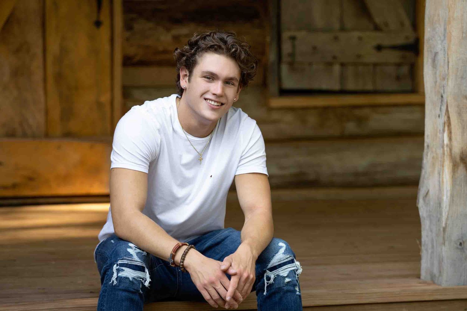 Young man in white t-shirt and jeans sitting casually on wooden floor with warm lighting in background.