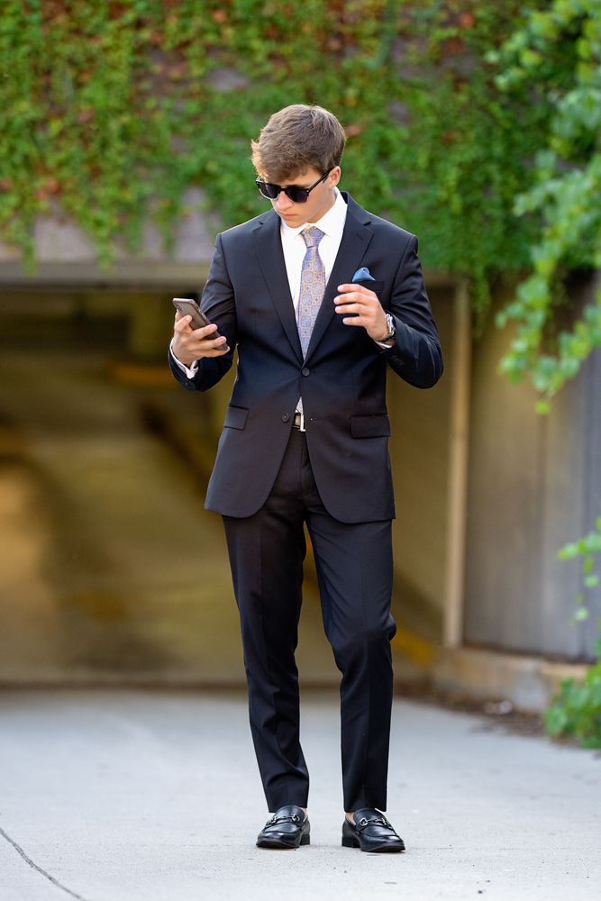 Person in formal black suit and sunglasses checks phone while standing outdoors.