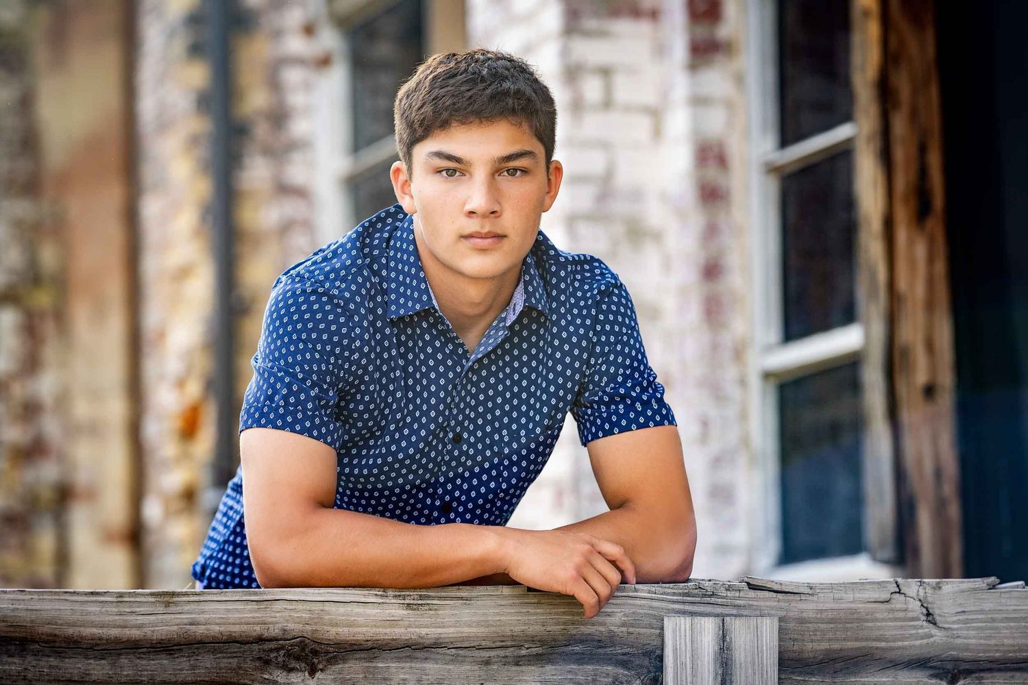 A person wearing a blue polka dot polo shirt leans against a rustic wooden railing near a brick wall.
