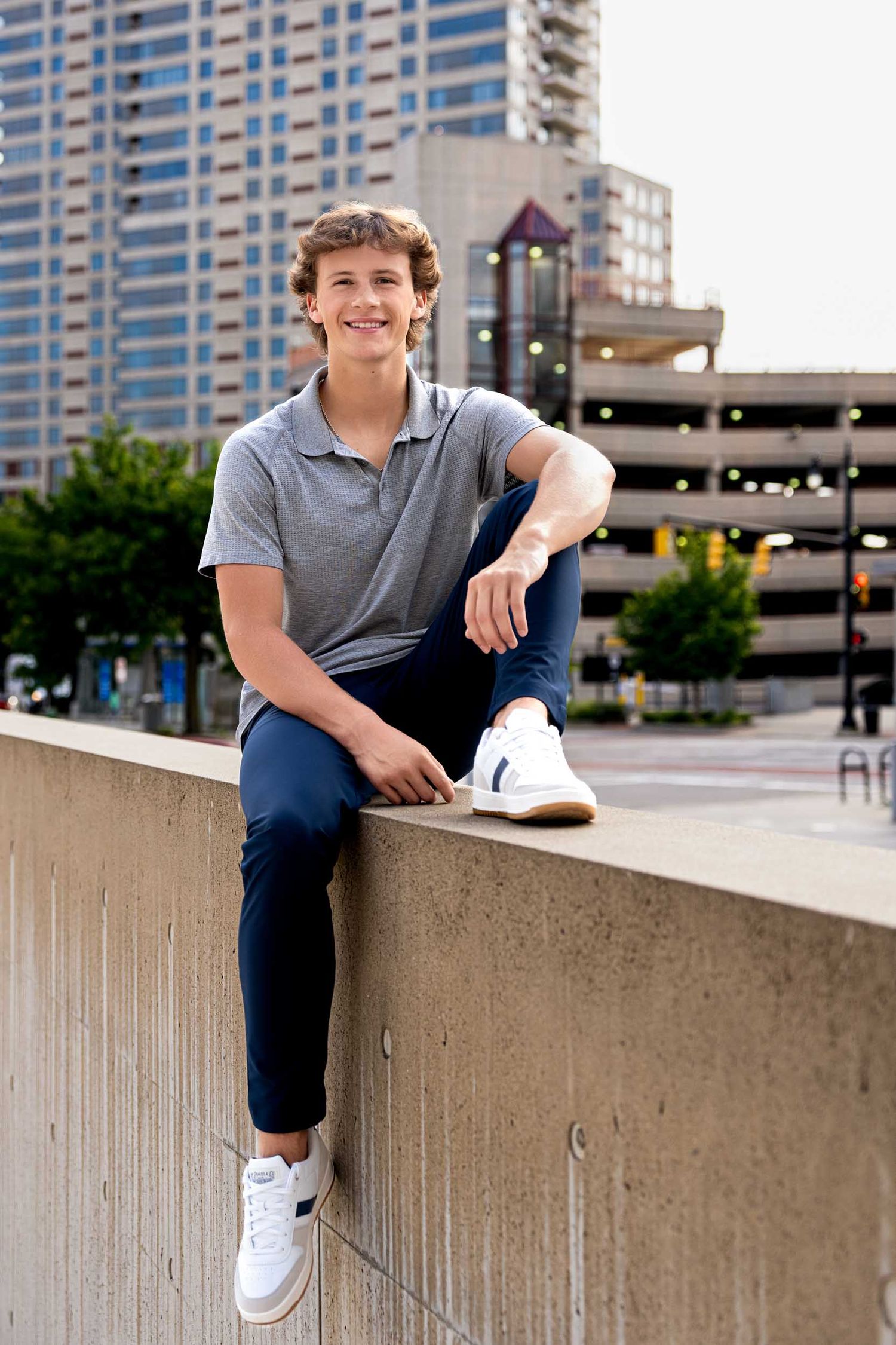Urban senior portrait session with person in grey polo and dark pants sitting on concrete wall.