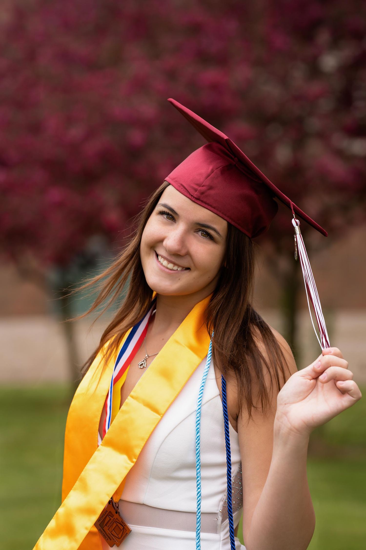 Graduate in red cap and gold stole poses with diploma against pink flowering trees in spring.